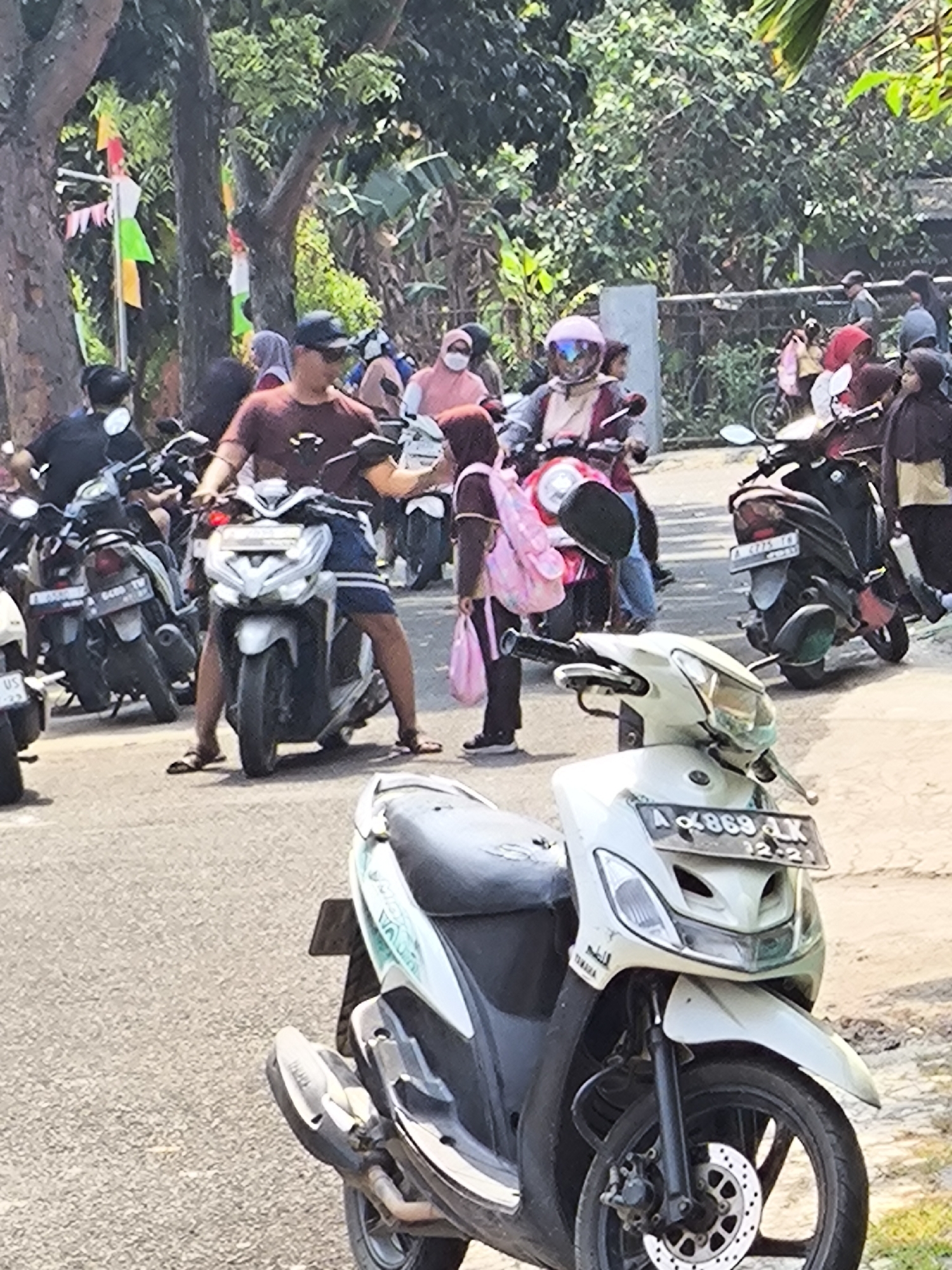 The image depicts an outdoor scene with several people on motorcycles, suggesting a traffic area or parking lot. The people are mostly young adults and some children, dressed in casual clothing and some wearing headscarves. They are interacting with each other, some looking around, others appearing to be waiting. Several motorcycles are parked or in use, with some riders preparing to depart or having just arrived. The environment is a paved road with trees and foliage in the background, indicating an urban or semi-urban setting. Decorative flags or banners, possibly for a local event or celebration, are visible, adding a festive element to the scene. The presence of multiple motorcycles and people suggests a communal gathering or a busy thoroughfare within the city of Cilegon, Indonesia.