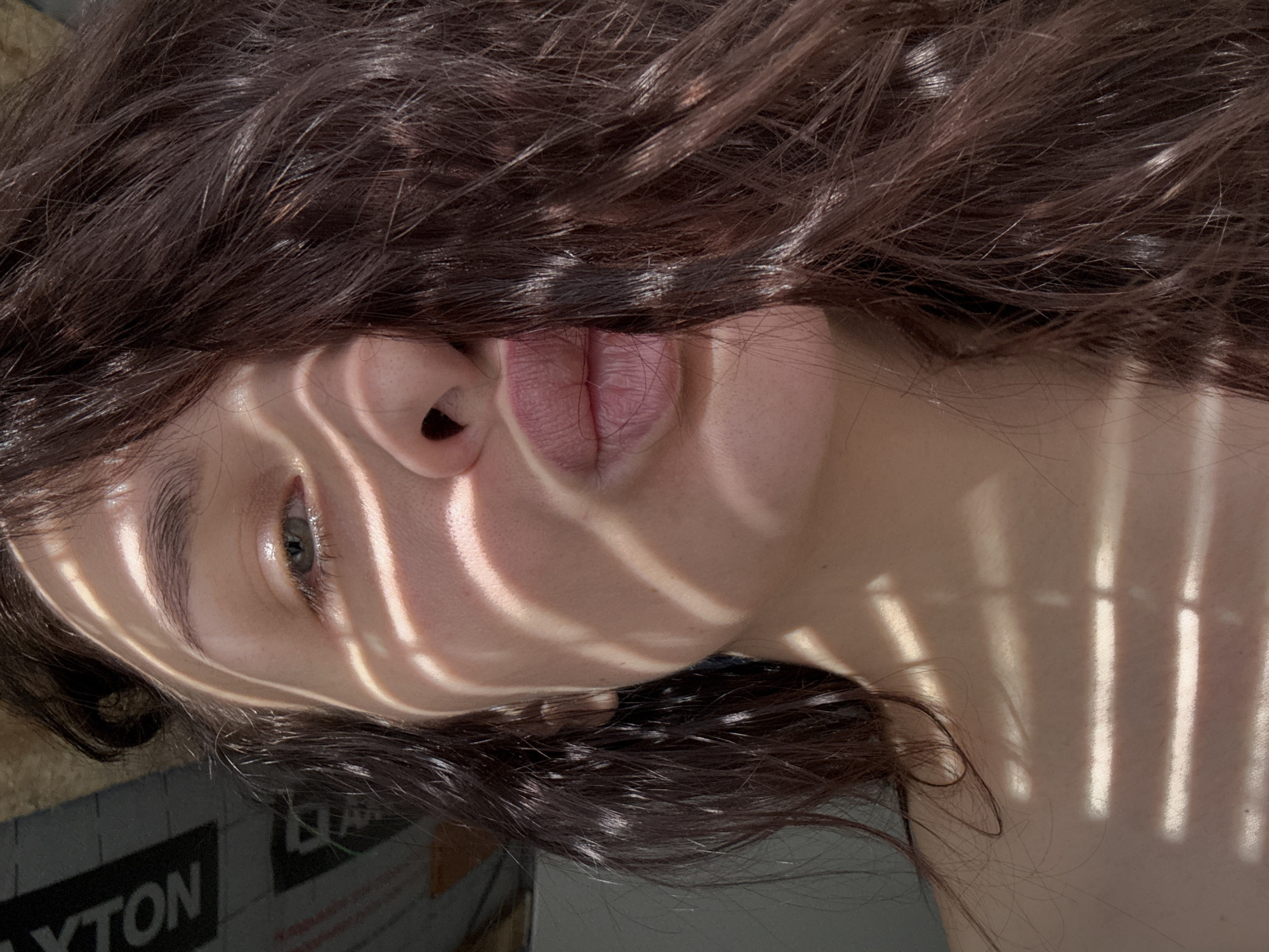 A close-up photograph depicts a female subject indoors, identified as being in Chaikovsky, Russia. Her left eye, light in color, is visible, along with her nose and mouth, which are pursed in a 'kissy face' gesture. Dark brown, wavy hair covers the right side of her face and extends over her bare left shoulder. Strong directional light creates prominent horizontal striped shadows across her face, neck, and shoulder, indicative of light passing through a slatted blind or similar aperture. A dark background element in the upper-left features faint, reversed text, appearing as 'NOLXYT'.