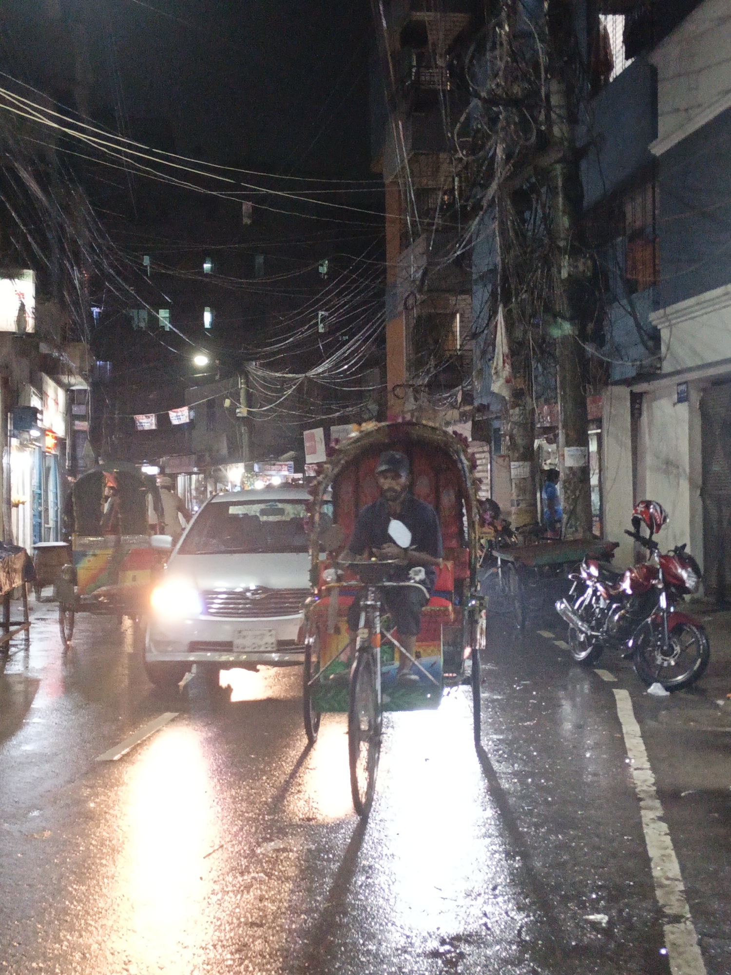 The photograph depicts a street scene at night, likely after rainfall, in the city of Unknown, Unknown. The pavement is wet and reflective, illuminated by headlights and streetlights. A man is operating a rickshaw, with the vehicle positioned prominently in the foreground. A white car with headlights on is visible behind the rickshaw. Two motorbikes are parked on the right side of the frame, with one appearing to have a helmet on the handlebars. Buildings line the street, with numerous overhead wires creating a dense network above. The lighting is generally dim, but with bright highlights from the vehicle lights. Other rickshaws and pedestrians are seen in the background.