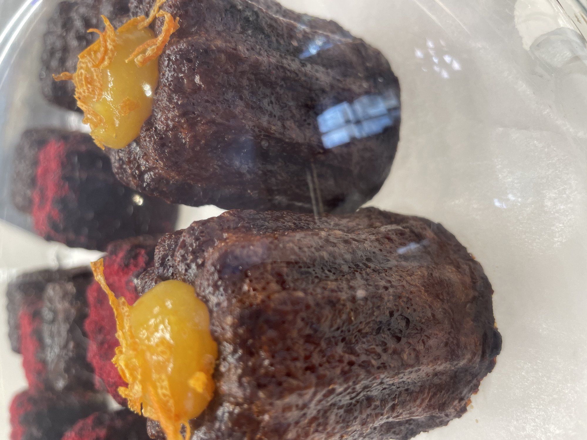 The image shows a close-up of several cannelés, a French pastry, displayed in a glass case. The cannelés are dark brown, with a distinct ridged texture and a slightly caramelized, almost burnt exterior. Two of the cannelés in the foreground are topped with a dollop of bright yellow filling, garnished with what appear to be thin strands of orange zest. In the background, other cannelés are visible, some with a dusting of red on top. The display case is clean, and the lighting suggests an indoor environment, likely a bakery or patisserie. There are no people or specific landmarks visible in the image that would allow for identification of a location in Belgrade, Serbia.