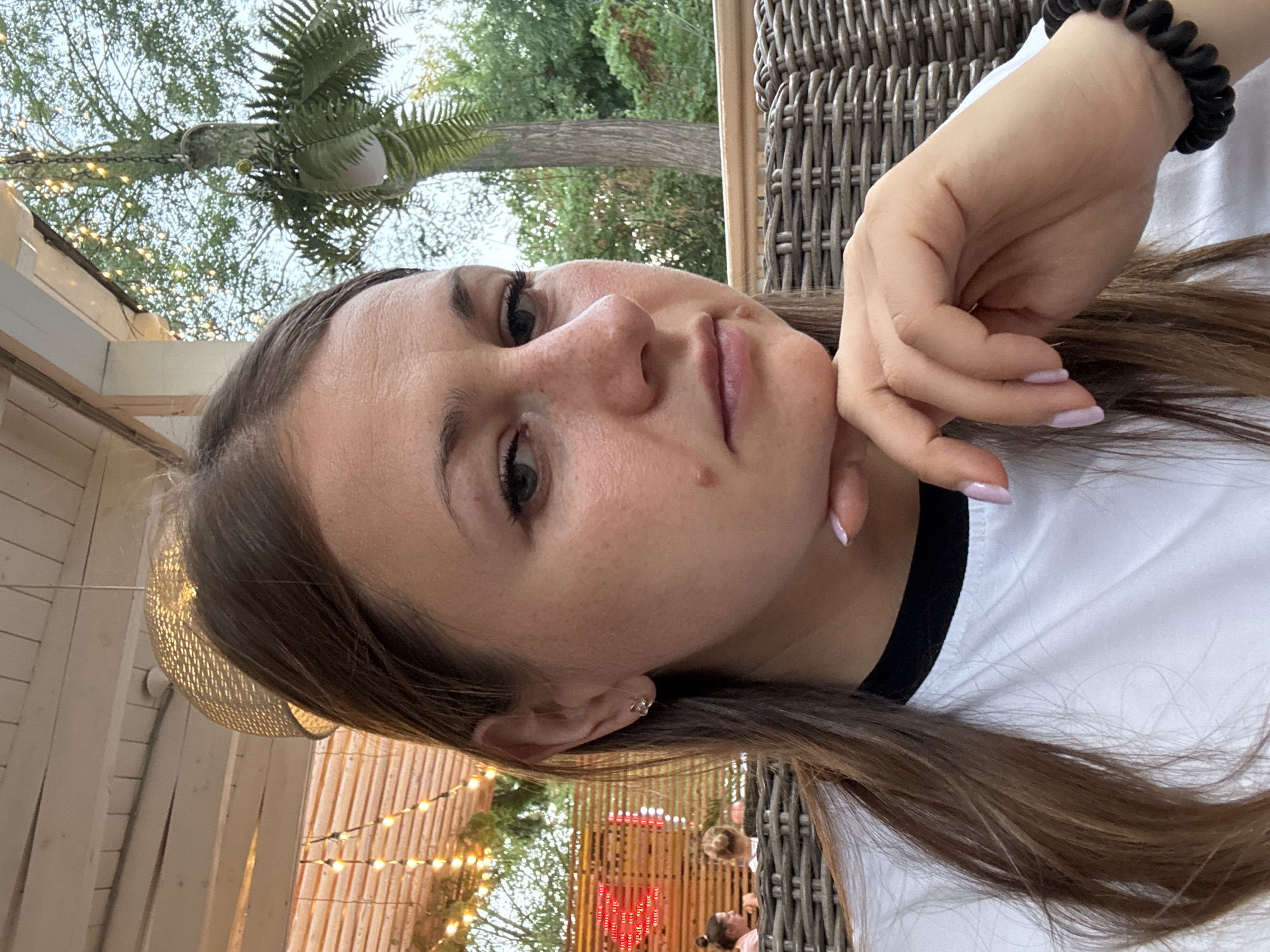 A medium close-up portrait captures a young woman with long brown hair and a thoughtful expression, her chin resting on her hand. She is seated outdoors at what appears to be a cafe or restaurant with wicker furniture. In the background, string lights are visible, strung across a wooden structure, and beyond that, trees and foliage suggest a natural, outdoor setting. The woman is wearing a white t-shirt with a black collar and a black coiled bracelet on her right wrist. Her fingernails are painted a light pink. The environment suggests a relaxed, possibly evening atmosphere.