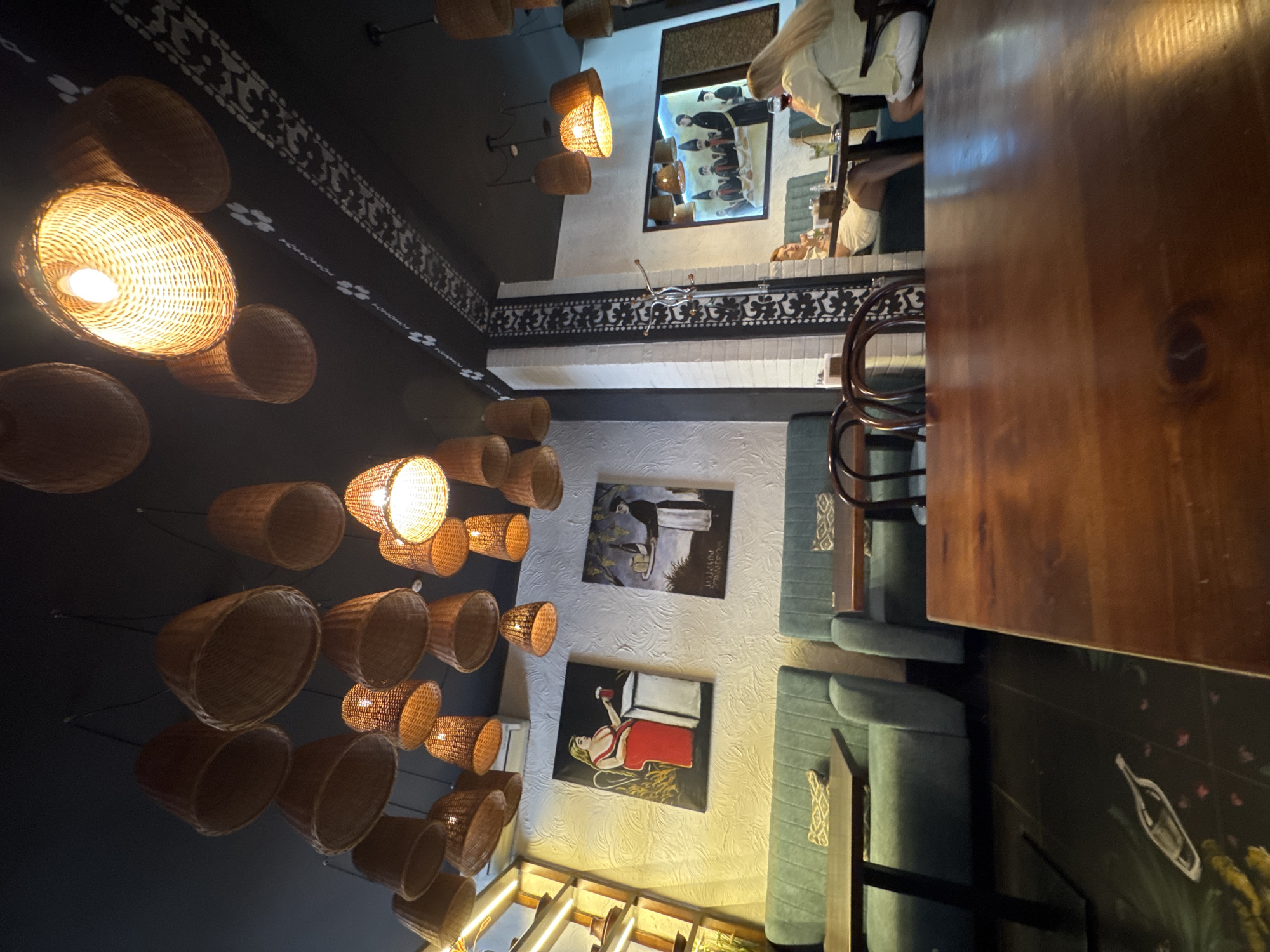 The photograph depicts the interior of a restaurant in Chernivtsi, Ukraine. The scene is illuminated by numerous hanging basket-like lamps with a warm, incandescent glow, casting light on the dark ceiling which features a decorative white pattern. In the foreground, a wooden table occupies a significant portion of the frame. Further back, a seating area with teal-colored couches is visible, each with a decorative pillow. On the wall behind the couches, two paintings are displayed: one of a woman in a red dress holding a drink, and another of a man carrying a tray with grapes. To the right, a framed picture shows several figures seated around a table. Two women are seated at tables in the background, one on the left and another on the right, both appearing to be patrons. The floor is dark and features a stylized image of a bottle and other decorative elements.