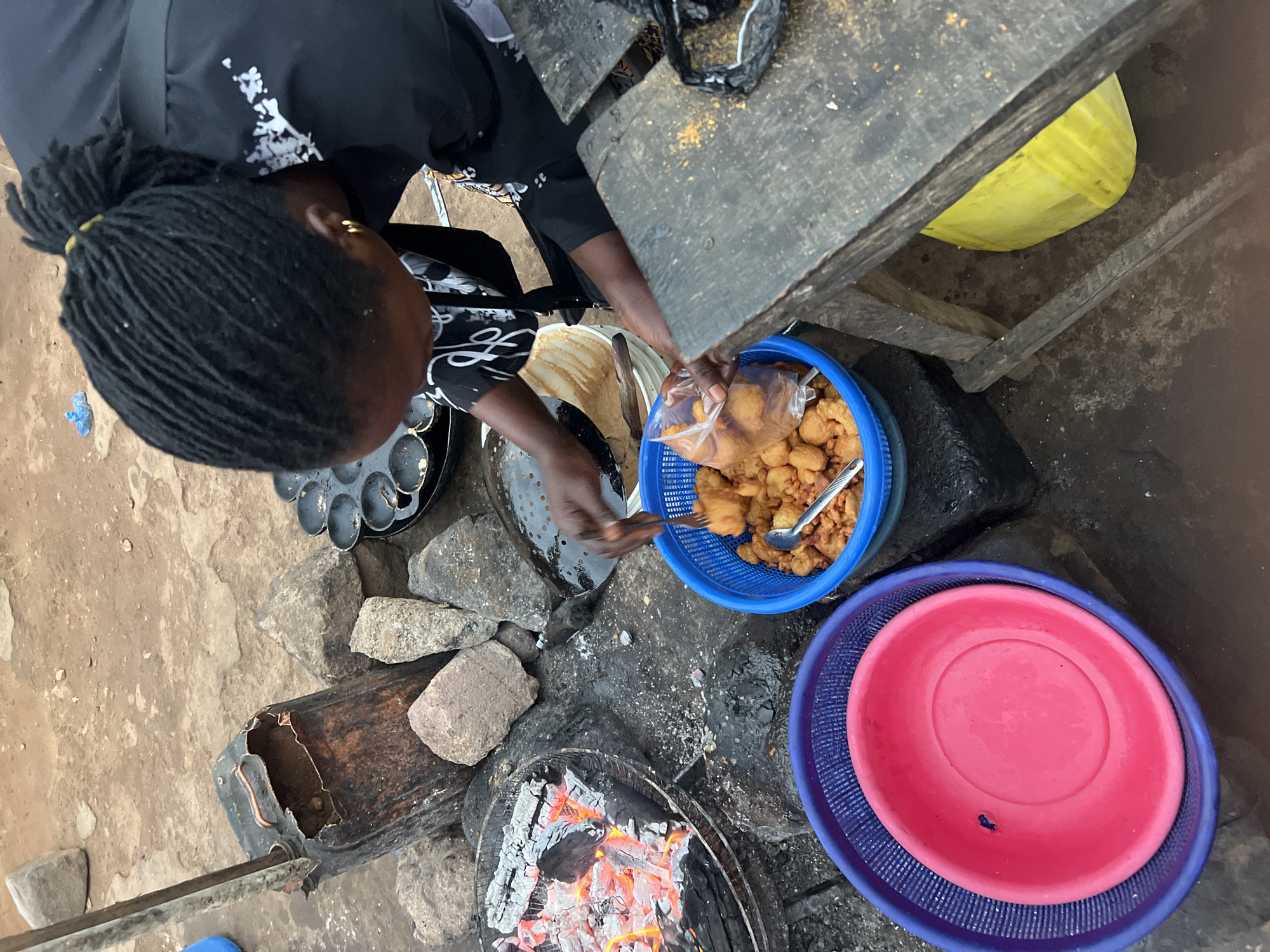 The image provides a top-down view of a woman engaged in outdoor cooking in Gura Top, Nigeria. The woman, wearing a black top with white patterns and her hair in dreadlocks tied back, is leaning over a cooking setup. Gold earrings are visible on her ears.
At the focal point is a charcoal stove with glowing embers, indicating active cooking. Several large stones are arranged on the unpaved ground, supporting the cooking implements. To the woman's left, a black metal pan with multiple spherical depressions, commonly used for frying items like 'akara' or 'puff-puff', is visible. Her hands are actively involved in the process, with one hand appearing to hold a utensil or tongs.
Adjacent to the cooking area, a blue plastic basket is filled with numerous golden-brown fried dough balls, some of which are already packed into a clear plastic bag. A spoon rests among the fried items. A bright pink plastic bowl is nested within a blue plastic strainer/basket on the ground.
A rustic wooden table or stand is partially visible in the upper right, with a yellow bucket placed underneath it. Near the charcoal stove, a broken brown plastic jerrycan lies on the ground. The scene depicts a bustling, traditional food preparation setting, likely a street food vendor, during daytime hours. No visible text is present.
