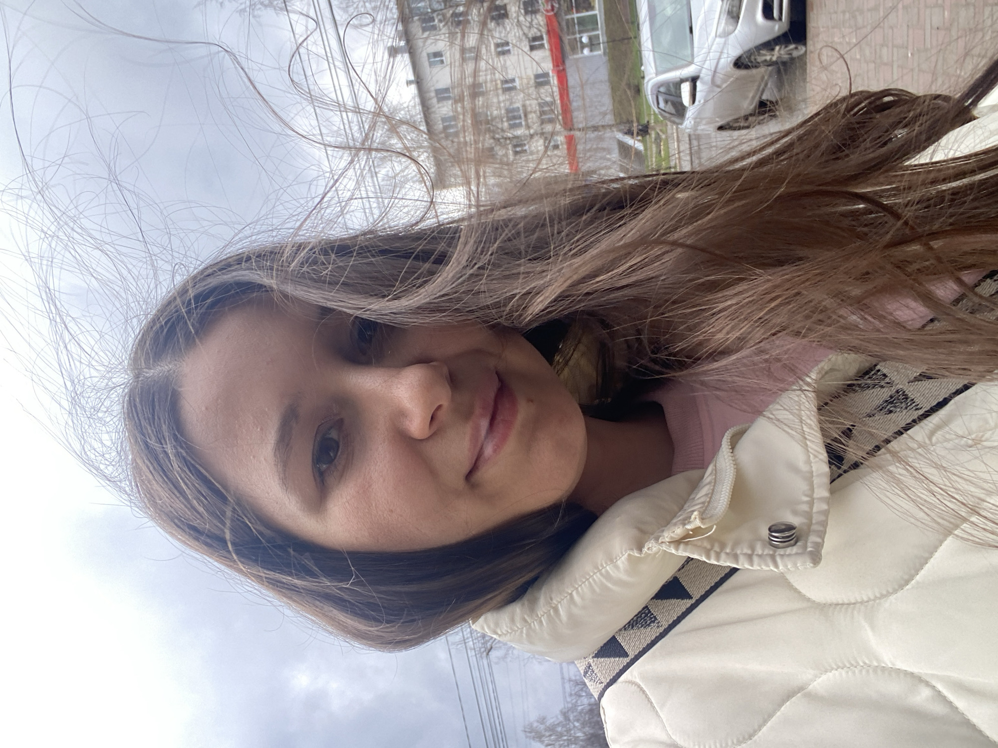 A young woman with long brown hair is standing outdoors on an overcast day. Her hair is blown by the wind. She is wearing a light-colored, quilted jacket over a pink sweater. In the background are apartment buildings and a white car. Powerlines are visible in the sky. Based on the available information, the location is the city of Chaikovsky, Russia.