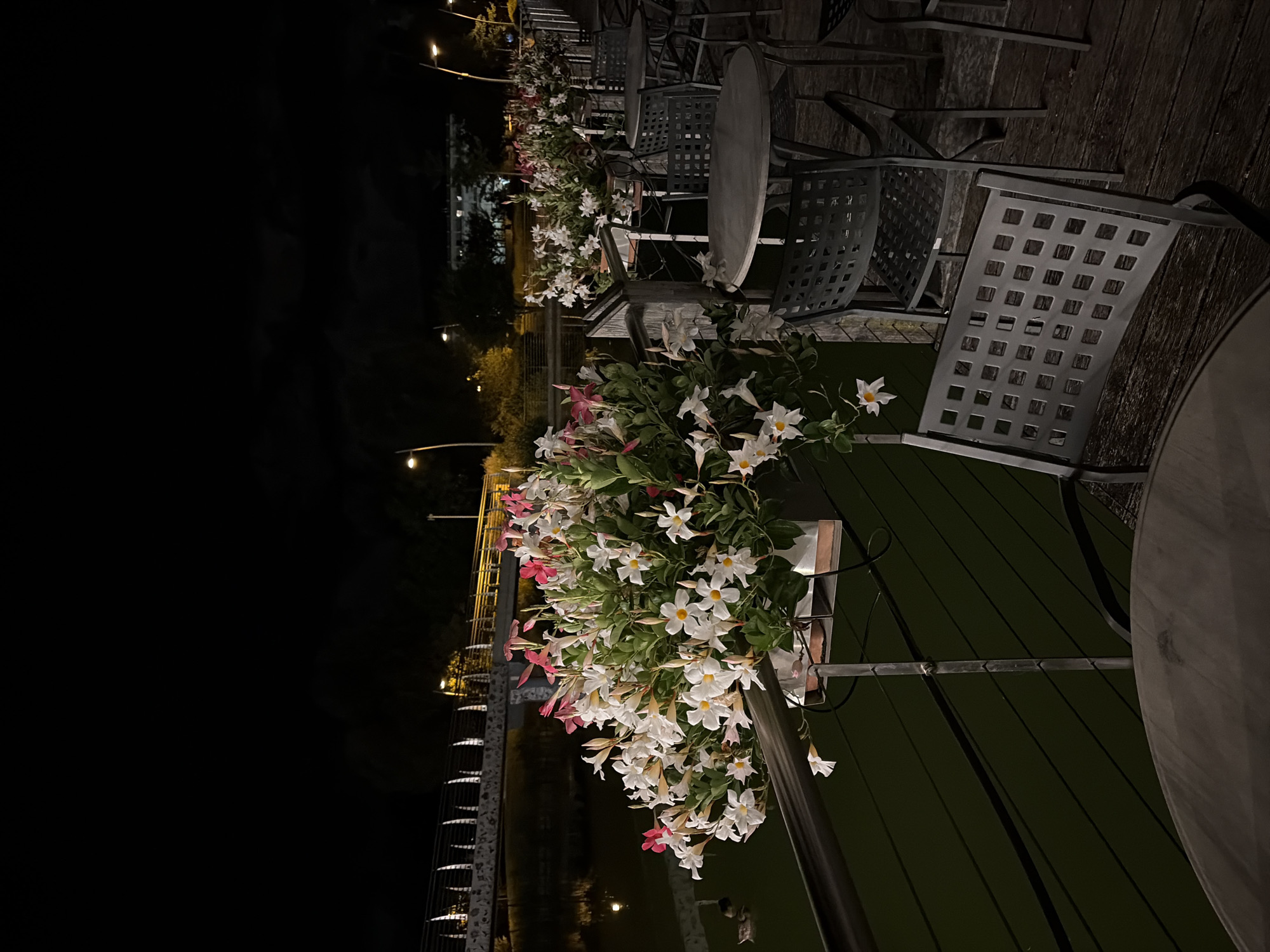 This is a photograph taken at night, likely from an outdoor dining area. The foreground features two round, dark-colored tables and several metal chairs with a grid pattern. In the midground, a railing runs horizontally, with a planter box filled with white and pink flowers cascading over it. Behind the railing, a dark body of water is visible, with a bridge illuminated by lights spanning across it in the background. The sky is dark, with some clouds present. The scene appears to be tranquil and possibly romantic, with the gentle lighting highlighting the flowers and the water. The location is not specified in the prompt.