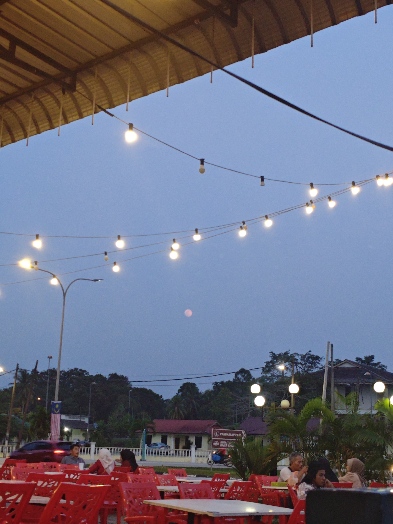 The image depicts an outdoor seating area with red plastic chairs and tables, likely a restaurant or cafe, in Kota Tinggi, Malaysia. The sky is a dusky blue, suggesting it is either early evening or late afternoon. A string of lights with visible bulbs is strung across the scene, some illuminated. In the background, there are trees, buildings with red roofs, a street light, and a distant full moon. A Malaysian flag is visible hanging from a pole. Several people are seated at tables, engaged in conversations or looking away. A red car is partially visible on a road in the mid-ground. The overall atmosphere is relaxed and casual.