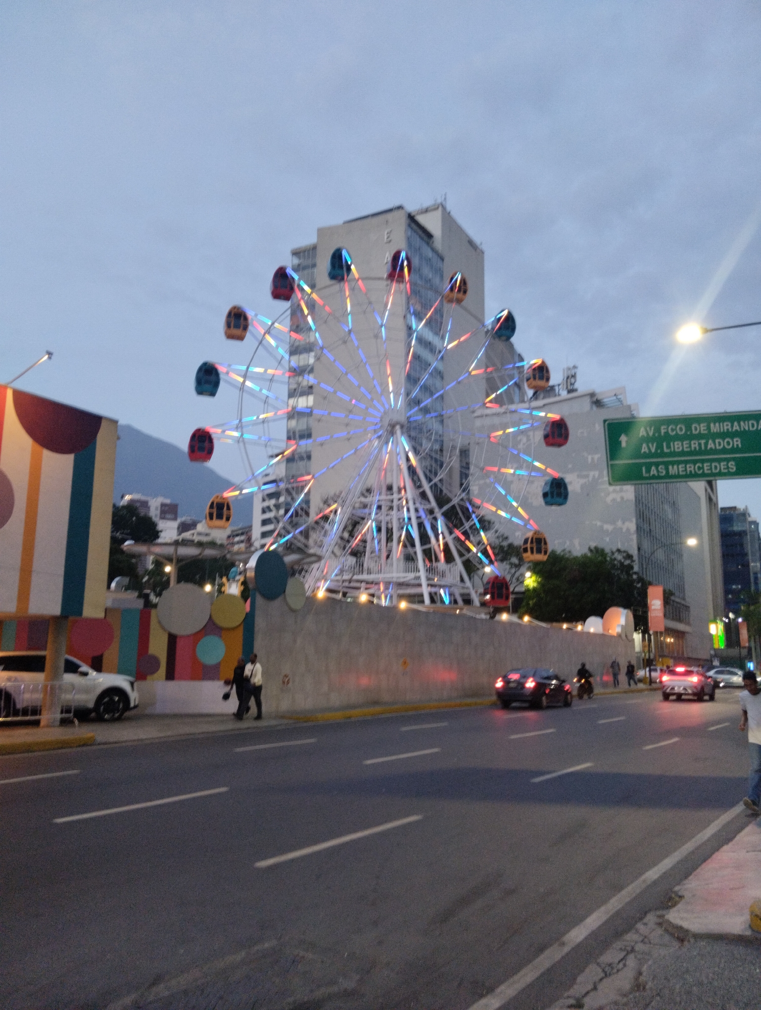 The image depicts a Ferris wheel illuminated with colorful lights in Caracas, Venezuela. The wheel is situated near a busy street, indicated by passing cars and pedestrians. A large building serves as a backdrop, and street signs for Av. Fco. de Miranda, Av. Libertador, and Las Mercedes are visible. A decorated wall with geometric shapes and a parked SUV are on the left side of the frame. Two individuals are walking along the sidewalk. The scene is captured during twilight, with a muted sky in the background.