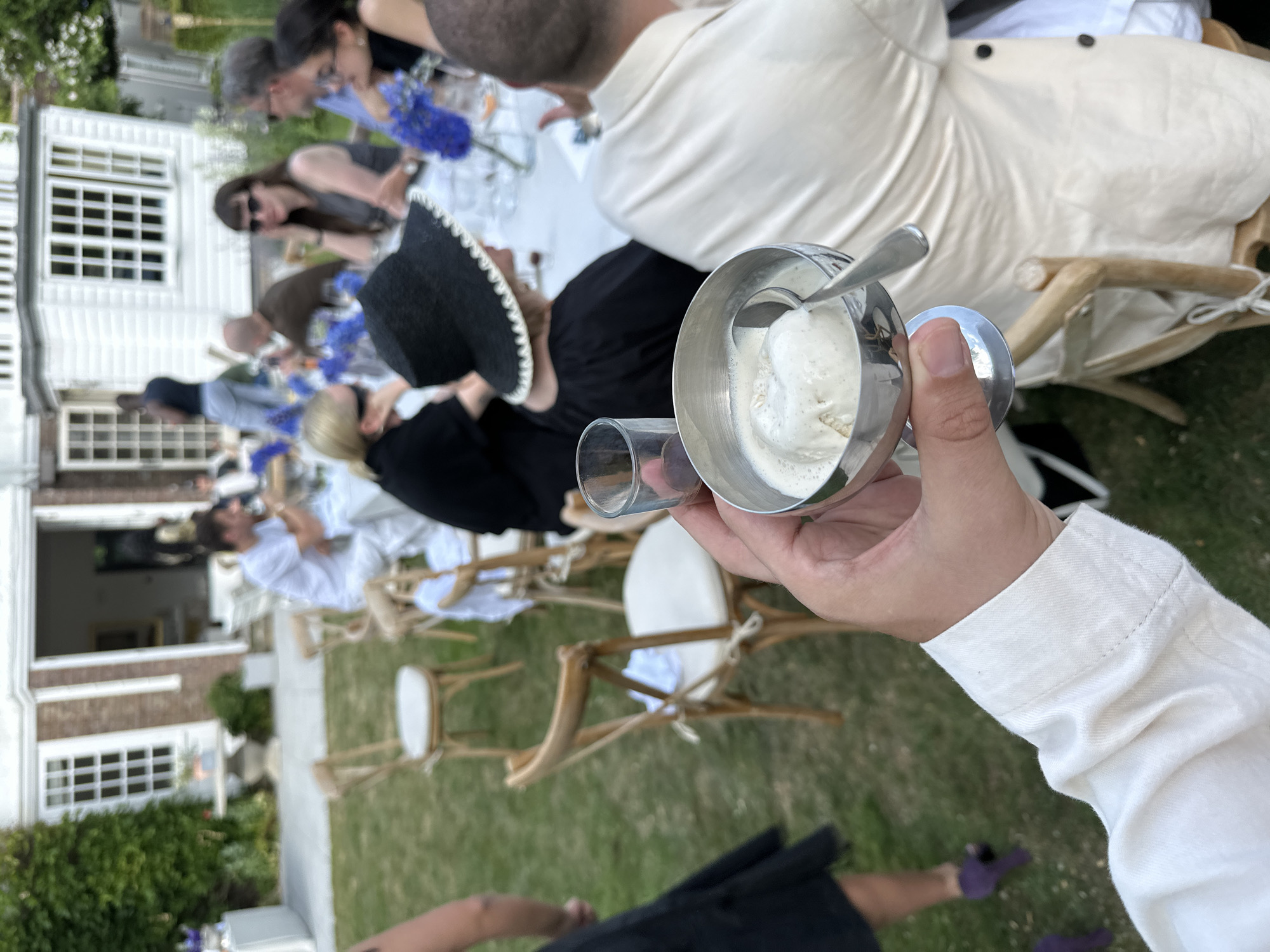 A person in a white shirt is holding a small silver cup of ice cream in their left hand and an empty glass in their right hand. The scene is outdoors, likely a garden or lawn. There are several other people seated at tables, some of whom are facing away from the camera. There are flowers on the tables. The setting appears to be a wedding or other celebratory event, with white chairs and tables in the background. A large building is visible in the background. This image does not contain enough evidence to confirm it was taken in the city of Cambridge, United Kingdom.