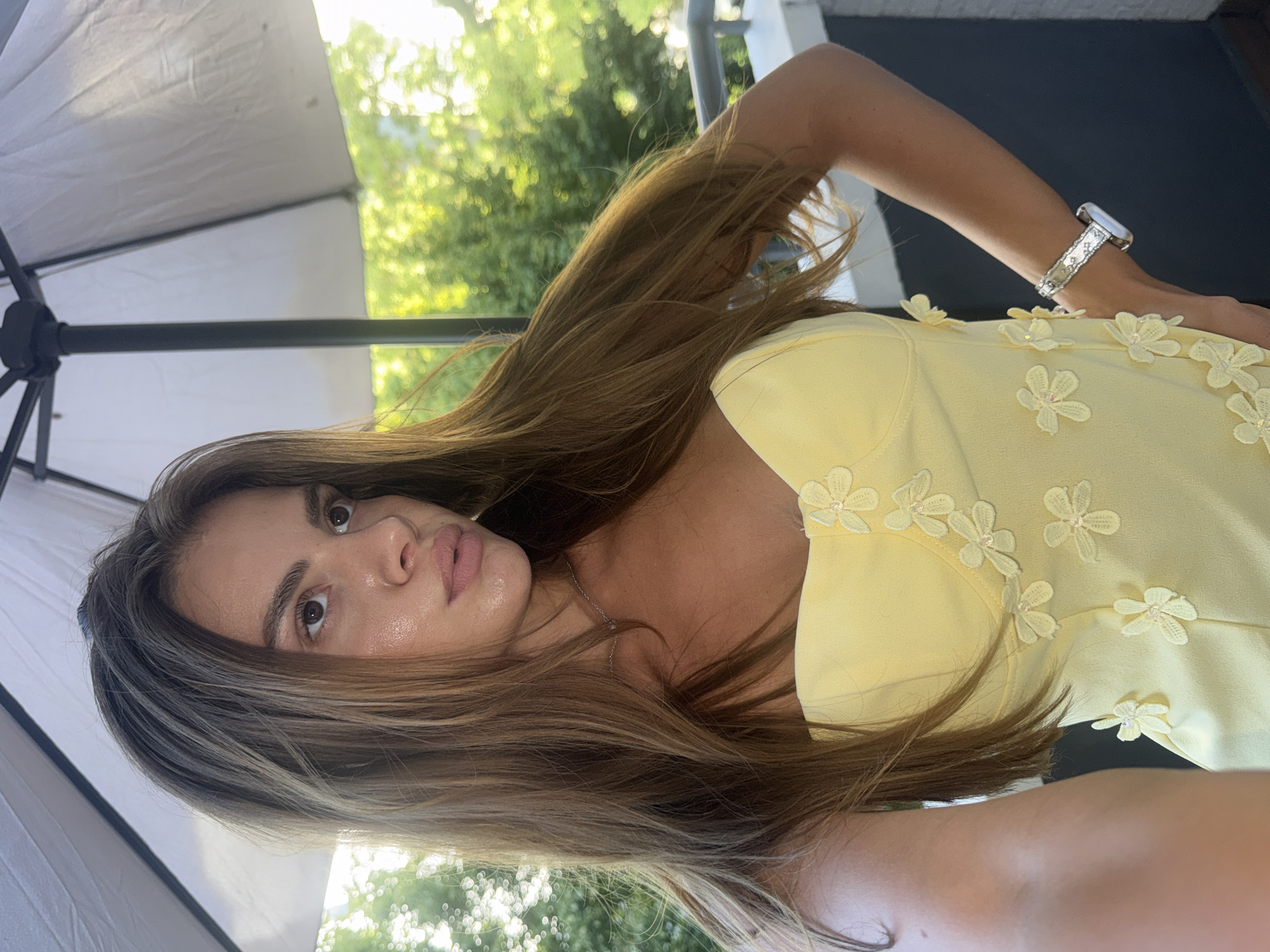 A young woman with long, brown hair is pictured from the chest up, wearing a light yellow strapless dress adorned with light yellow flower appliques. She is looking to her right, with her left hand on her hip and her right arm bent, her hand resting on her chest. A silver watch is visible on her right wrist. The background consists of lush green trees and a white umbrella. The scene appears to be outdoors, possibly on a balcony or terrace, given the railing visible in the lower right corner. The lighting suggests it is daytime, with sunlight filtering through the trees. There is no indication of the location being in Plovdiv, Bulgaria.