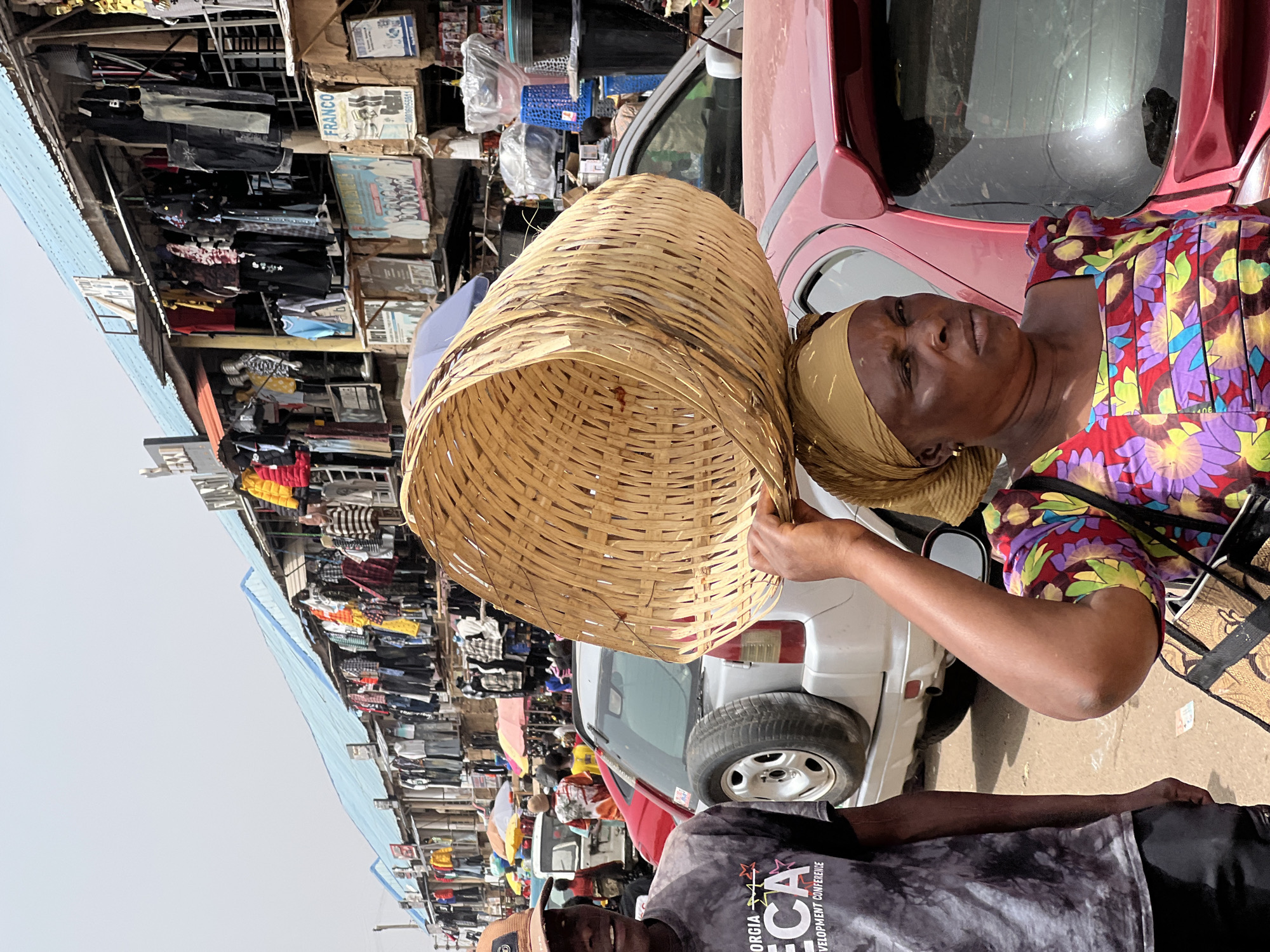 The image depicts a woman holding a large, woven basket in what appears to be a bustling marketplace in Bukuru, Nigeria. The marketplace displays various goods, primarily clothing items, hanging from stalls and buildings. A red car and a white SUV are parked nearby, indicating vehicular traffic within or adjacent to the market area. A man in a printed t-shirt is also visible in the foreground. The scene suggests a busy commercial environment.