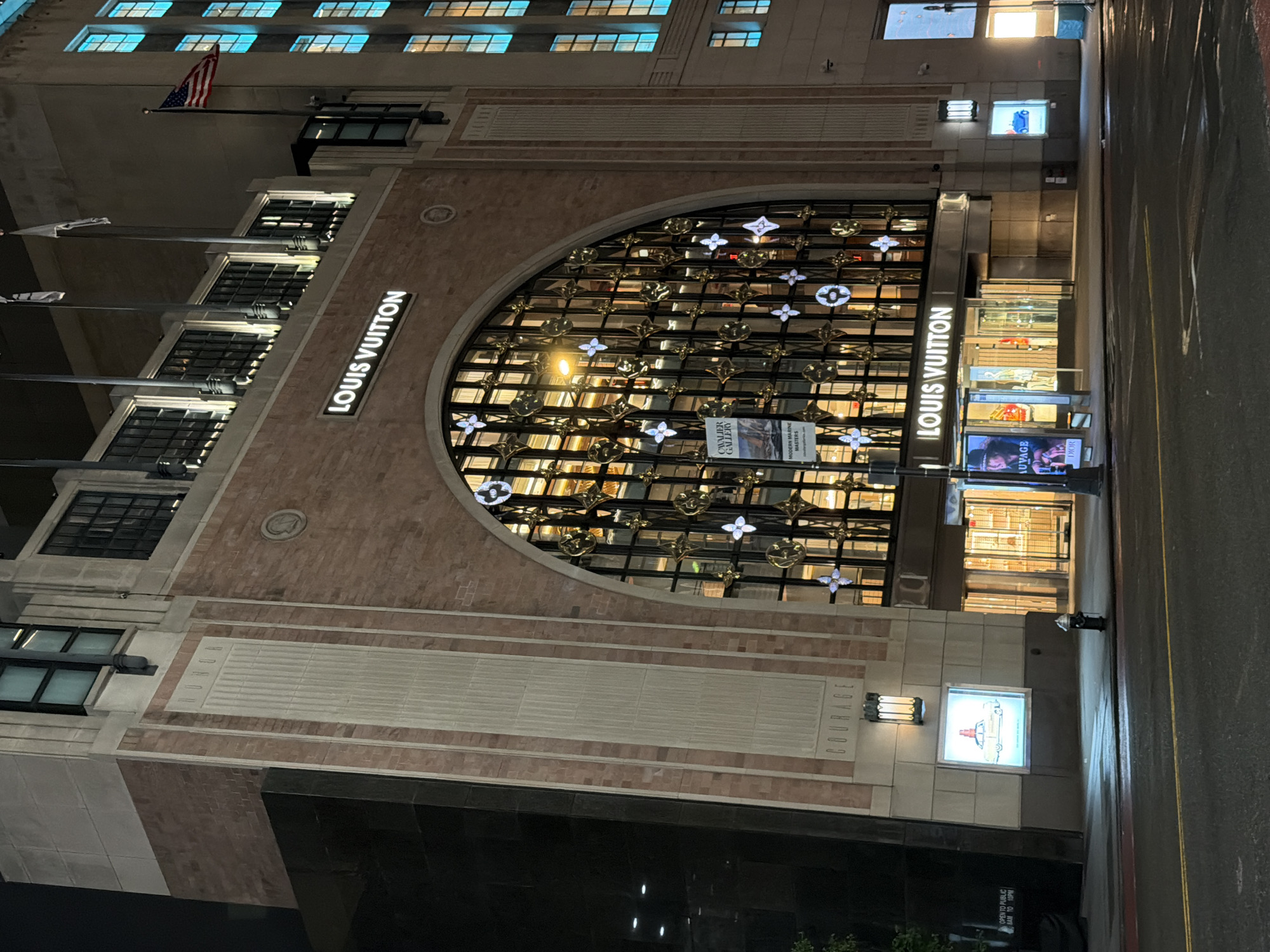 This is a nighttime exterior shot of the Louis Vuitton store located at 1 East 57th Street in New York City, New York, United States. The building features a large arched window with intricate gold and white patterned grillwork. The Louis Vuitton logo is prominently displayed in illuminated white lettering above the arched window. The facade is made of brick and has vertical stone elements. To the left of the store entrance, a large poster features a black and white image of a woman. To the right of the entrance, a large digital display shows an image of a blue car. The street in front of the store is paved, with yellow and white road markings visible. A fire hydrant stands on the sidewalk. The windows on the upper floors of the building are illuminated with a blue glow. An American flag is visible on a flagpole to the right of the building.