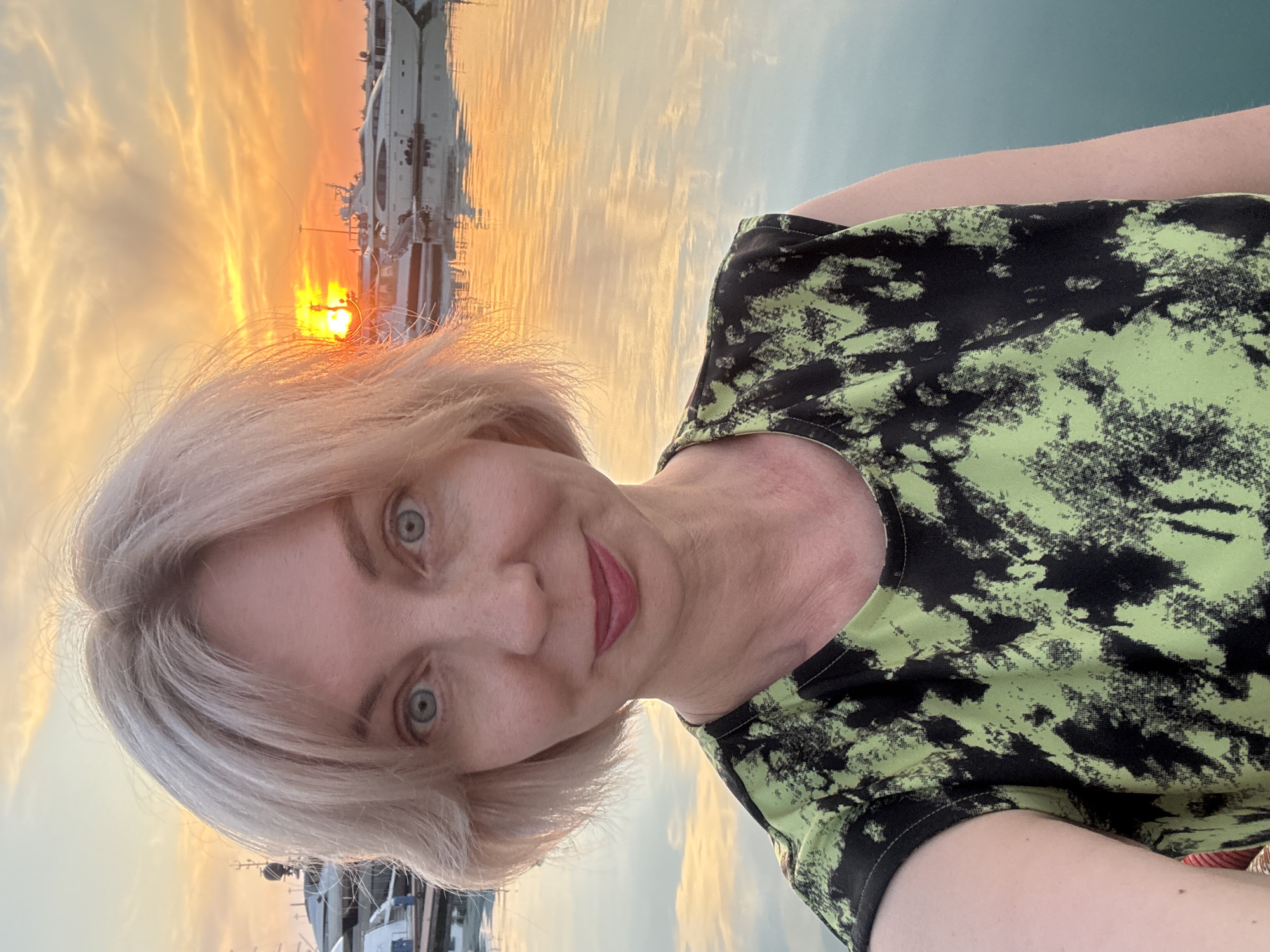 An individual selfie photograph captures a fair-skinned female subject with short, light blonde hair and blue-grey eyes, appearing to be in her late 40s to early 60s. She is facing the camera with a direct gaze, exhibiting a slight smirk with crimson-colored lips, and is wearing a sleeveless top featuring an abstract lime green and black pattern. The image is taken at sunset in a marina located in Sochi, Russia. The sun is visible as an orange orb near the horizon on the right side, casting a warm, golden light across the scene. The sky displays hues of orange, yellow, and pale blue, with scattered clouds. The calm water in the foreground and midground reflects the sunset colors and the surrounding boats. Several large white yachts and smaller vessels are docked or moored in the background, with their reflections visible on the water's surface. Dark-colored dock structures are also present on the left side of the frame.