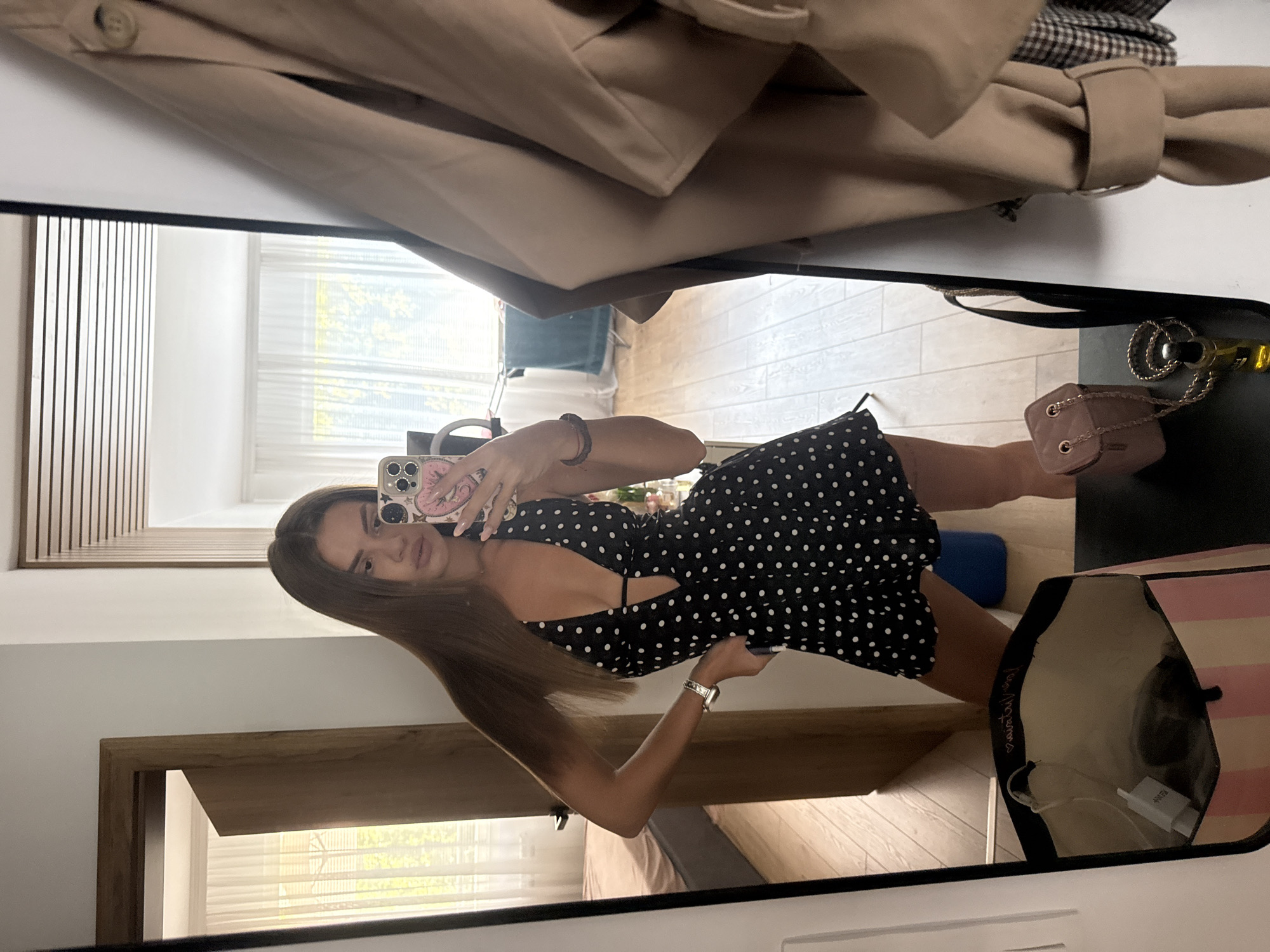 A young woman stands in front of a full-length mirror, taking a selfie. She is wearing a black, short, polka-dot dress with a deep V-neck. Her hair is long and brown, and she is wearing a watch on her left wrist and a bracelet on her right. A pink phone case with cartoon characters is visible in her hand.
The mirror reflects a room with light-colored wooden flooring and white walls. To the left, a doorway leads to another room where a bed is visible. White curtains cover a window in the background, allowing natural light to illuminate the space. A beige trench coat hangs on a hook to the right of the mirror. In the foreground, below the mirror, a pink and white striped bag and a small brown purse with a gold chain are placed on a surface. There is also a bottle of perfume. The scene appears to be indoors, possibly in a residential setting. There is no specific indication that the location is in Plovdiv, Bulgaria.