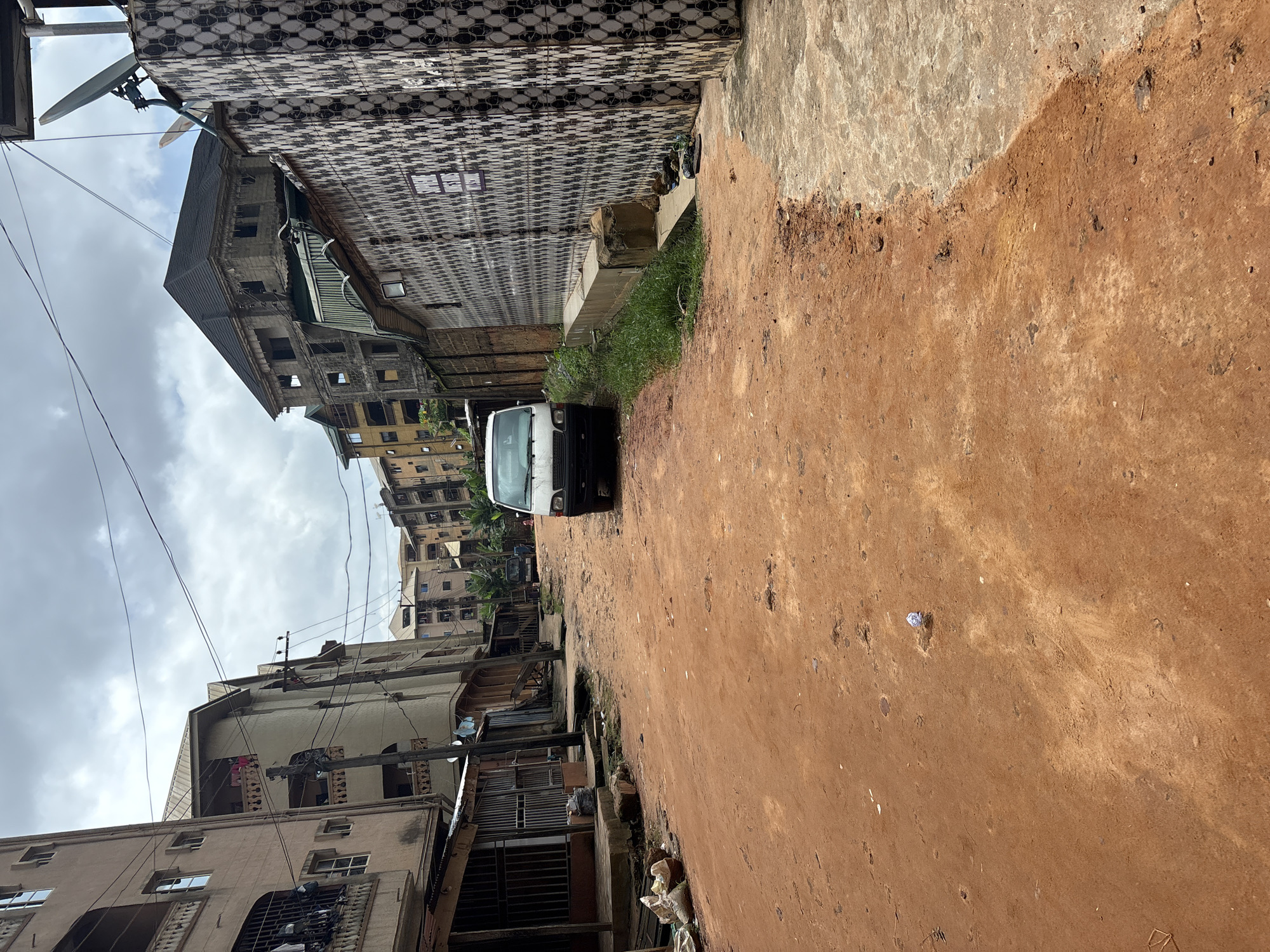 The image depicts a street scene in Onitsha, Nigeria. A dirt road, characteristic of many areas in the city, dominates the foreground, showing signs of wear and tear. Several multi-story buildings line the sides of the road, displaying a mix of architectural styles and varying states of upkeep. Some buildings appear newer and more maintained, while others show signs of age and neglect. A white van is parked on the right side of the road, facing away from the viewer. The street is relatively empty, with no people clearly visible. Overhead, power lines crisscross the sky, which is overcast with clouds. The overall impression is one of a busy urban environment with evident infrastructure challenges.