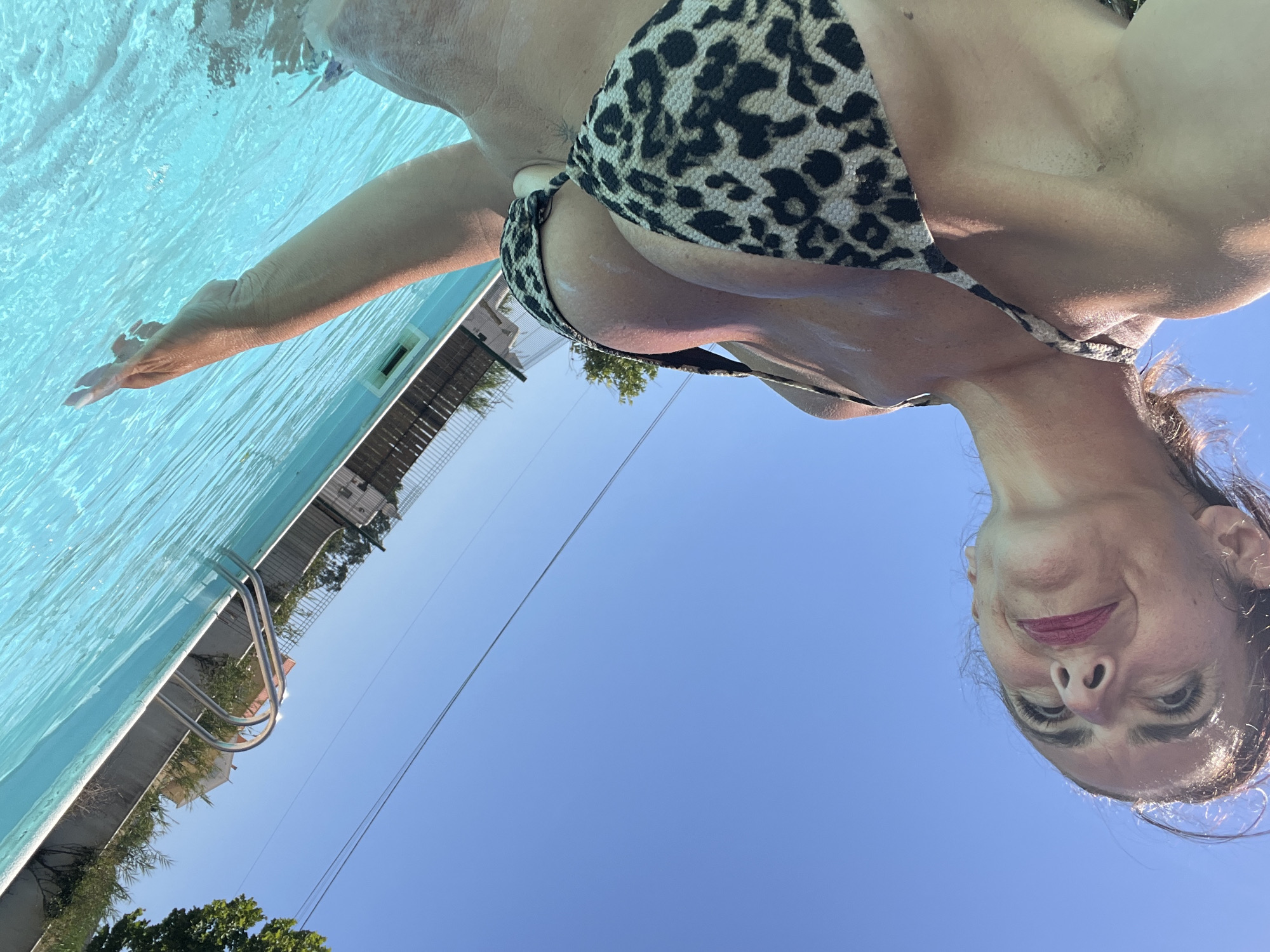 A low-angle, close-up shot captures a woman in a leopard-print bikini standing in a swimming pool under a clear blue sky. The woman's face is turned upwards and to the right, with a smile playing on her lips and a hint of makeup. Her wet hair is pulled back, and her arm is extended into the pool, her fingers just touching the turquoise water. The pool's edge and a portion of the surrounding fence and vegetation are visible in the background. The image is likely taken in Caldas da Rainha, Portugal, given the architecture and landscaping clues often found in that region.