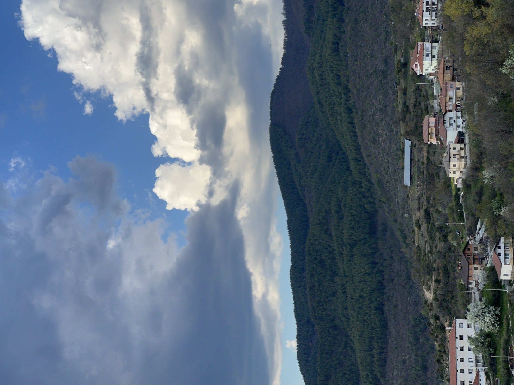 The image depicts a mountainous scene in Kribul, Bulgaria, under a sky with a mix of blue and heavy, grey clouds. A densely forested mountain stretches vertically in the middle of the frame, abutting the sky. Houses and structures are situated on the right side of the mountain, appearing as if built into the hillside. These buildings range in size and color, with some appearing white, others having red roofs, and all seemingly residential. The sky on the left is a mix of clear blue with some puffy white clouds and heavy grey clouds indicating potential rainfall.