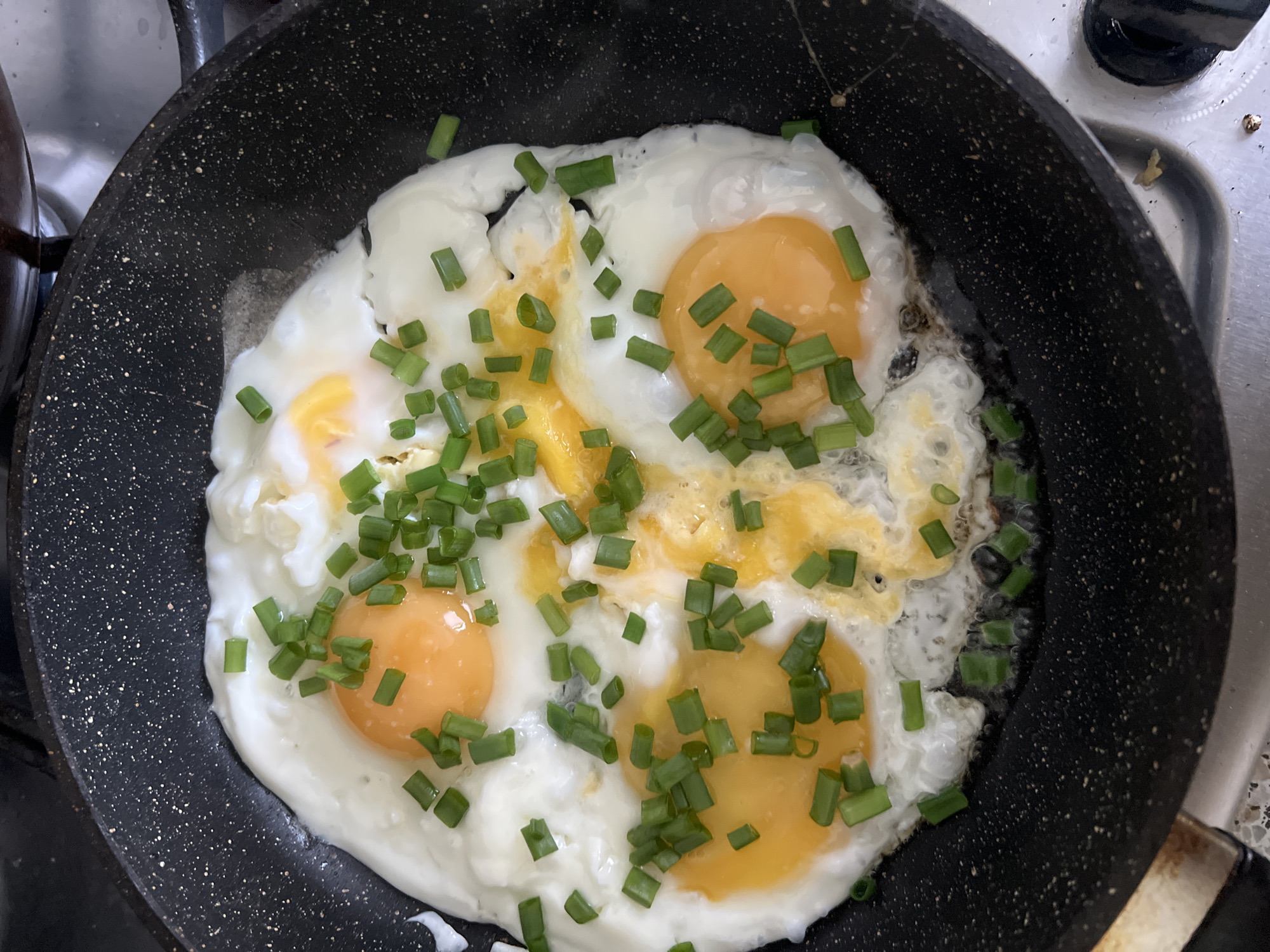 The image shows a black frying pan containing four fried eggs, sprinkled with chopped green onions. The pan is on a stovetop. The location is unknown but presumed to be in the city of Chaikovsky, Russia based on the user's query. There are no people visible in the image.
