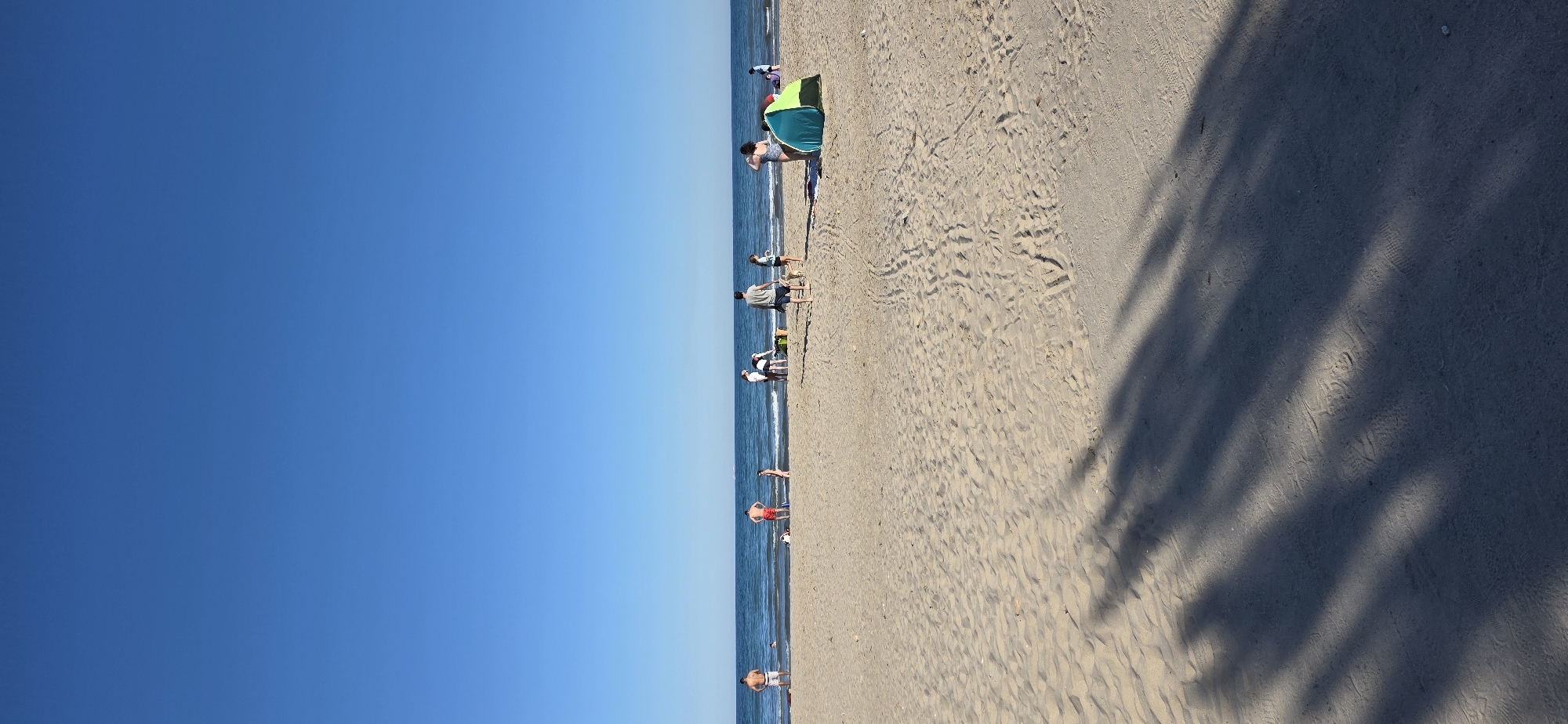 This is an outdoor photograph of a beach in Canet d'en Berenguer, Spain, taken on a sunny day with a clear blue sky. The photograph is oriented vertically, with the sky occupying the left half of the frame and the beach and sea on the right. The beach is sandy, with some areas showing patterns of footprints and wave marks. The sea is a deep blue, and there are several people in the water or near the shoreline. Some people are standing or walking along the shore, while others appear to be swimming or playing in the water. There is a green and blue beach tent set up on the sand. The right part of the photo has shadows from the trees.