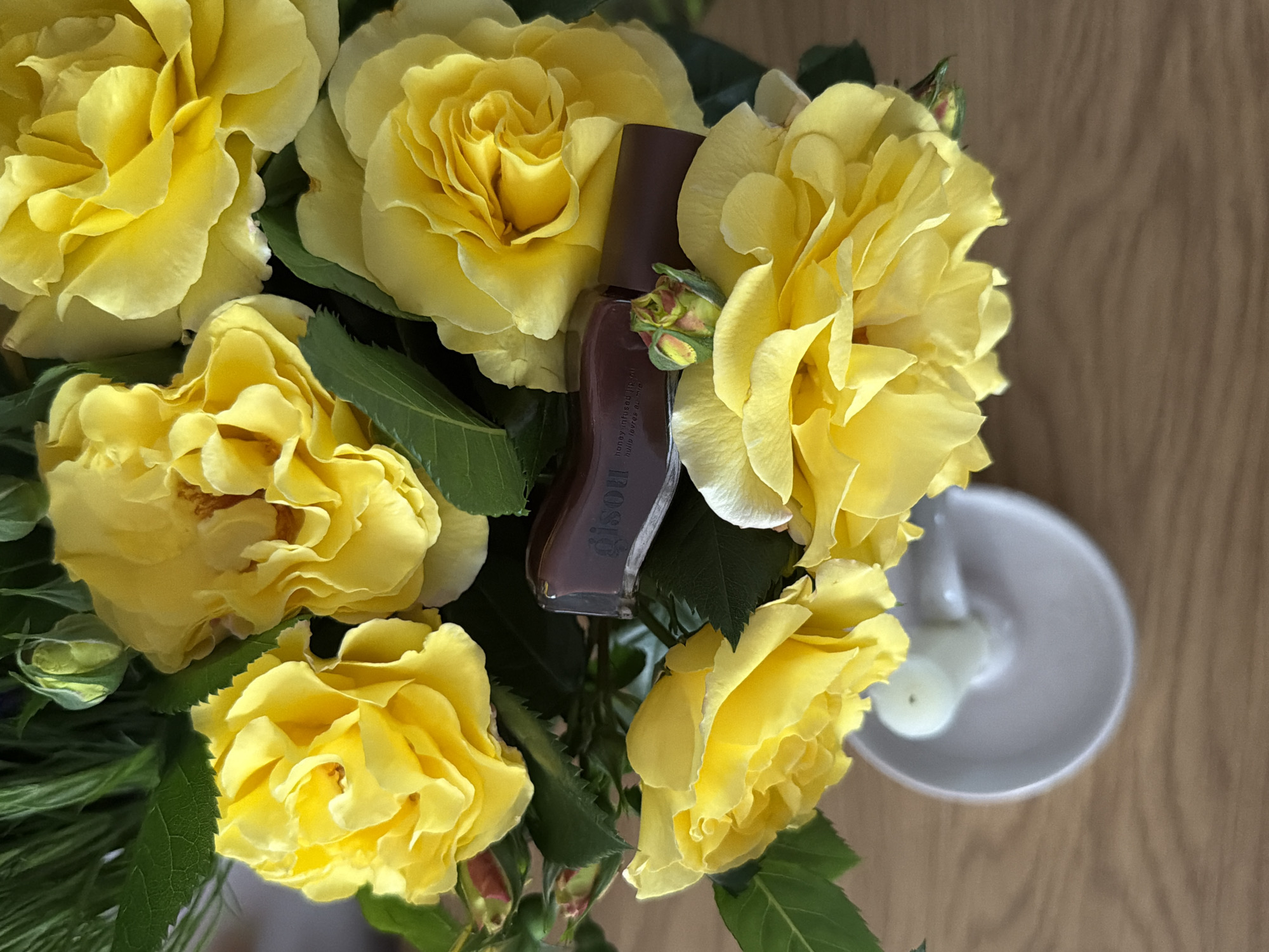 The image depicts a close-up of a bouquet of yellow roses on a wooden surface. Nestled amongst the roses is a bottle of "gisou" branded lip oil. To the right, partially obscured by the roses, is a small ceramic dish containing a white candle. The background is a neutral, light brown, suggesting a tabletop or other similar surface. The location details are not present in the image.