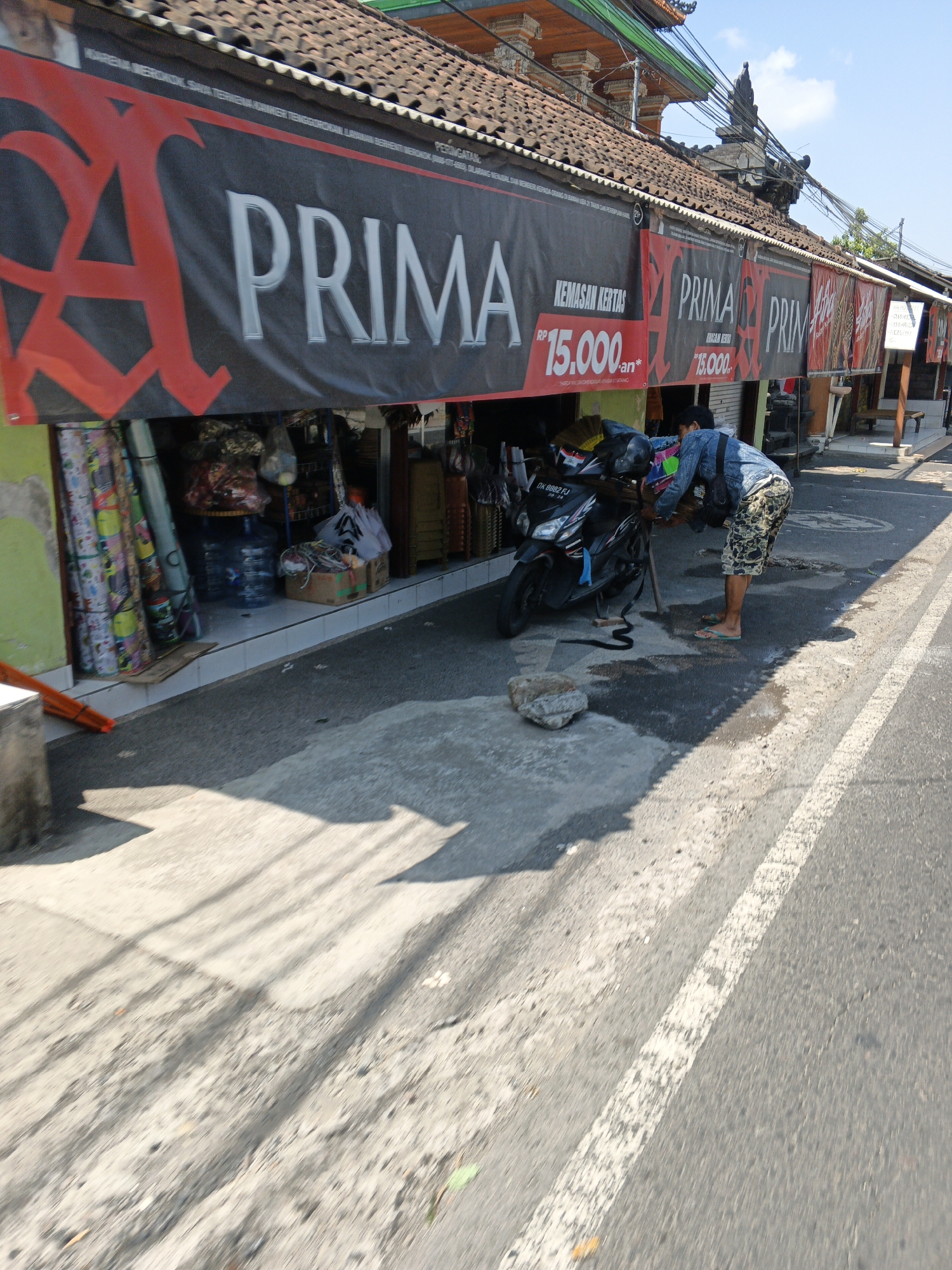 The image depicts a street scene in Ubud District, Indonesia, with a focus on a small shop or stall. The shop front features a large black banner with red lettering that reads "PRIMA" and displays prices in Indonesian Rupiah. Behind the banner, various goods are displayed, including what appear to be rolls of fabric or wallpaper, bottled water, and other items. In front of the shop, a black motorcycle is parked, and a person wearing a denim jacket and camouflage shorts is leaning over the motorcycle, appearing to adjust something. The ground in front of the shop is paved, with some water or wet patches. The street itself is asphalt, marked with a white line. The architecture visible suggests a tropical environment, with a tiled roof and decorative elements typical of Indonesian buildings. The overall impression is a bustling, everyday scene in a commercial area.