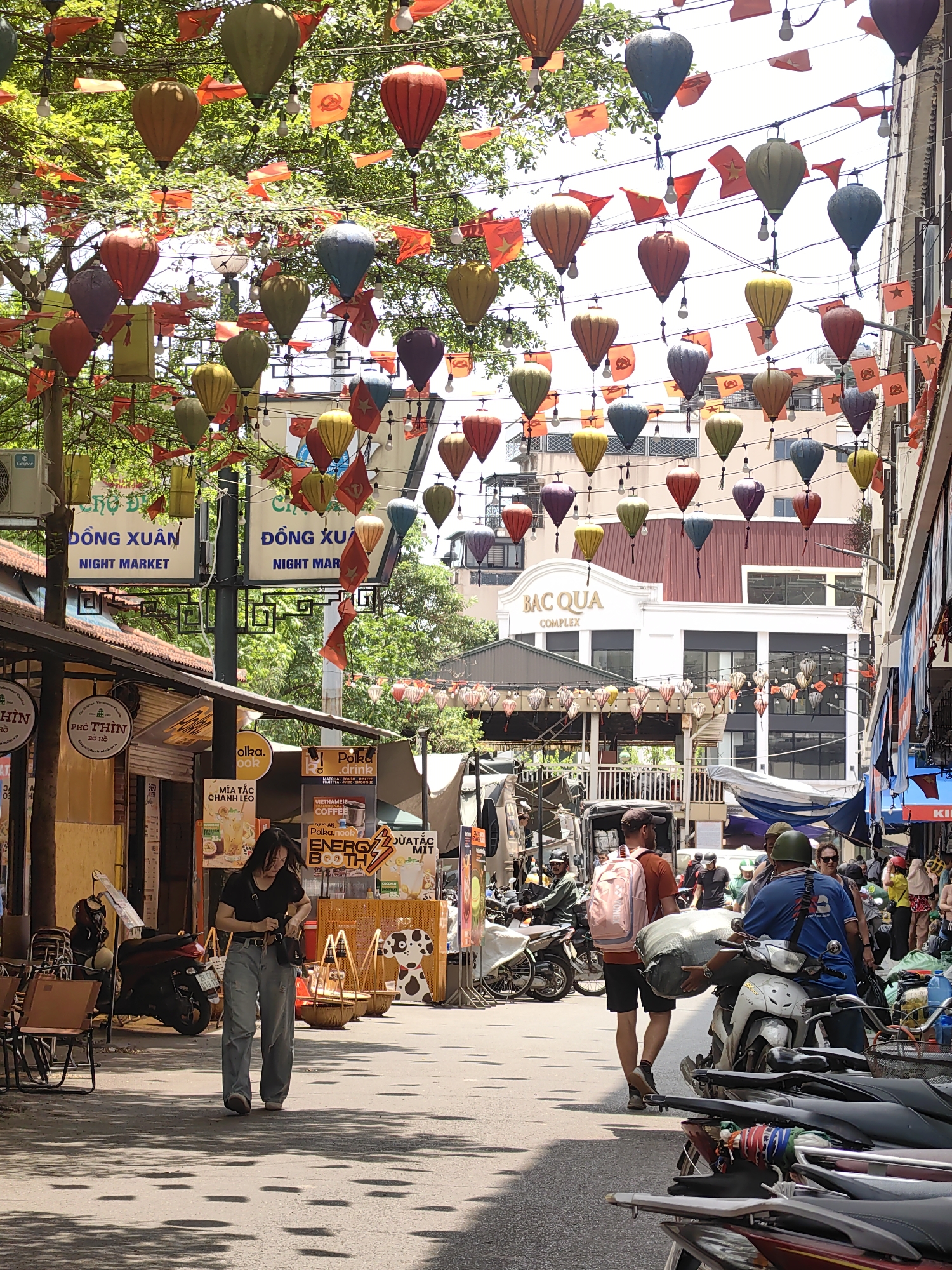 The image captures a lively street scene in Hà Nội, Vietnam, likely near the Đồng Xuân Night Market or a similar commercial district, suggested by signs for "ĐÔNG XUÂN NIGHT MARKET" and "BAC QUA COMPLEX." The narrow street is bustling with daily activity under a canopy of lush green trees, adorned with numerous colorful lanterns resembling hot air balloons and red rectangular flags featuring a yellow star. String lights are also woven through the festive overhead display.
The bright daylight indicates a sunny day, casting distinct shadows of the trees and decorations onto the paved ground. On the left, several open-air shops and food stalls line the street, with tables and chairs set out. One sign clearly reads "PHỞ THÌN BỜ HỒ," while another advertises "MÍA TẮC CHANH LEO." A visible "Casper" air conditioning unit is attached to a building.
Various people navigate the street. In the foreground, a woman wearing a black t-shirt and light blue jeans walks with a handbag, looking down. Further down the street, individuals are seen walking and riding motorbikes. A man with a pink backpack and an orange t-shirt walks beside a person on a white scooter, who is wearing a blue shirt and a green helmet, carrying a large sack. More motorbikes are parked along the right side of the street, indicating common urban transport. Shopfronts on the right display signs for beverages and food, including "Polka.drink," "VIETNAMESE COFFEE," "Polka.nook," and "ENERGY BOOTH," adding to the vibrant commercial atmosphere. The overall impression is one of a busy, cultural marketplace.