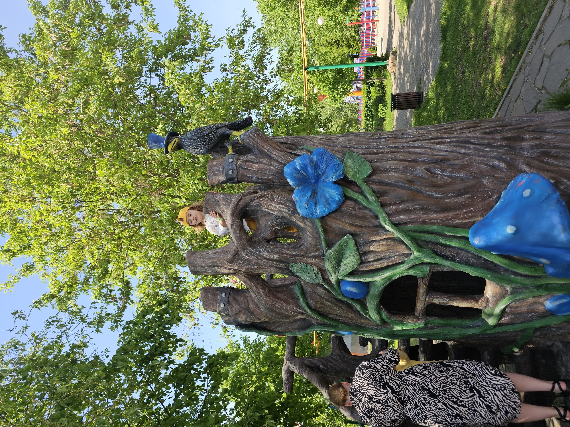 A vibrant outdoor scene unfolds in the city of Khimki, Russia. A sculpted tree trunk, painted in shades of brown with bright blue flowers and mushrooms, stands as a focal point. A child wearing a yellow hat peers from an opening in the tree trunk, while a woman in a black and white patterned dress stands nearby. Atop the tree, a sculpted bird adds to the whimsical atmosphere. The backdrop features lush green trees and a paved pathway, with glimpses of a colorful structure in the distance. A trash can is visible along the path. The sky above is a clear blue.