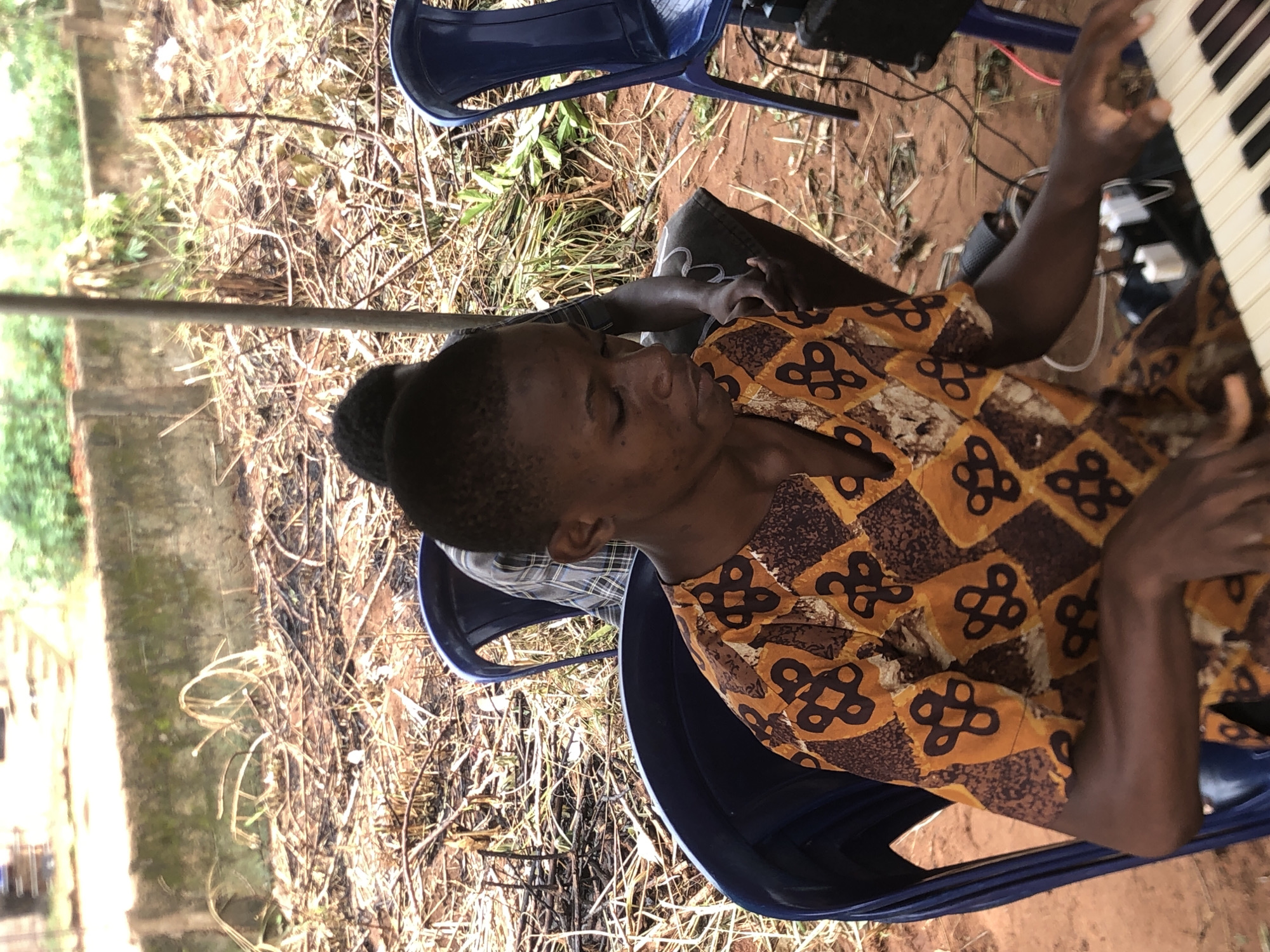 The image shows a person seated on a blue plastic chair, playing a keyboard. The person is wearing a patterned shirt with brown and orange motifs. The background consists of dry vegetation and bare ground, possibly indicating an outdoor setting in a rural or semi-rural area. The scene could potentially be located in the city of Ukwu-Abwa, Nigeria.