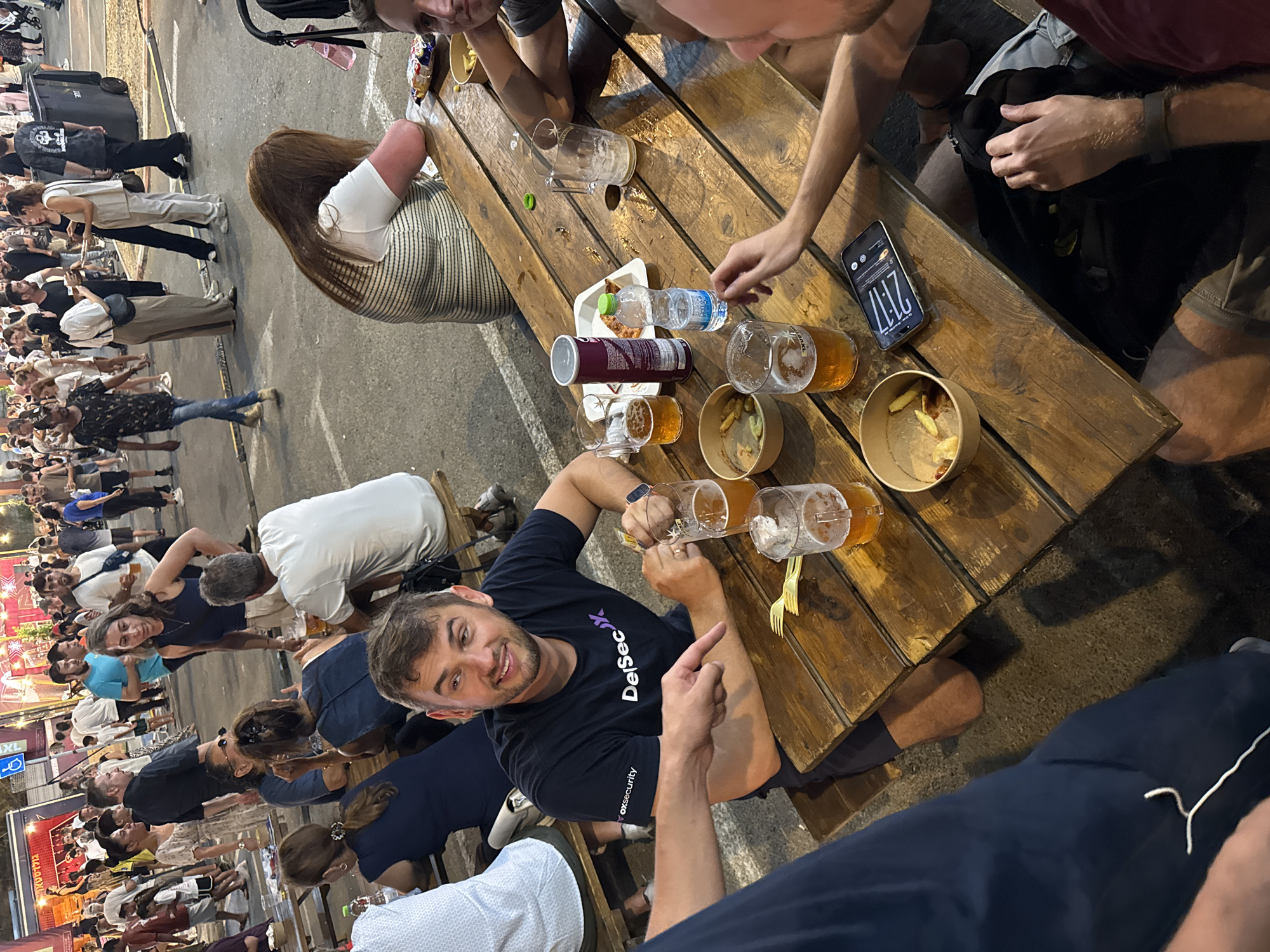 The image depicts a street scene in Netanya, Israel, with a group of people gathered around a long wooden picnic table. The table is laden with food and drinks, including glasses of beer, a bottle of water, a Pringles can, and small containers of snacks. Several individuals are seated at the table, engaged in conversation and enjoying their beverages.
In the foreground, a man wearing a black t-shirt with the "DelSecX" logo is prominently featured. He is smiling and looking to his right, appearing to interact with someone off-camera. To his left, another person's hand is visible, pointing towards him.
The background of the image shows a lively street environment, with more people walking around and stalls or kiosks lining the street. The overall atmosphere appears to be casual and social, possibly at an outdoor market or event. The lighting suggests it is evening.