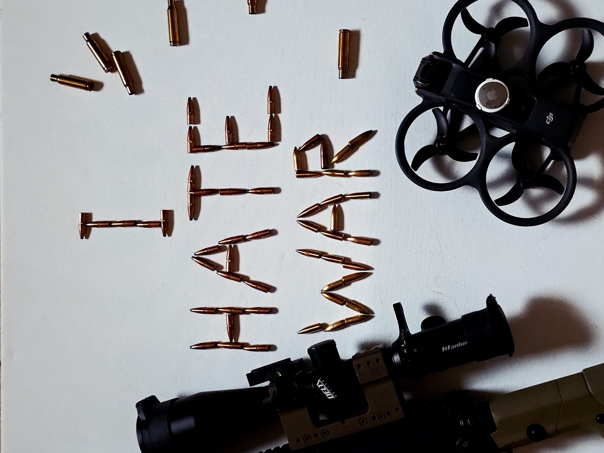 The image presents a symbolic still life composition on a plain, light-colored surface. The central focus is the phrase "NO WAR," meticulously spelled out in capital letters using numerous metallic, golden-hued bullet projectiles and brass cartridge casings. Loose casings are scattered in the upper left corner.
In the upper right, a black drone with propeller guards is visible, identifiable by the "DJI" logo on its side. A circular silver sticker featuring a bitten apple logo (resembling the Apple brand) is affixed to the top of the drone.
The bottom right of the image shows a section of a rifle. A scope, partially obscuring the word "DELTAPOINT" and clearly labeled "titanium," is mounted on the dark-colored firearm. The arrangement deliberately uses objects associated with conflict—bullets, a rifle, and a drone—to convey a strong anti-war message. The directional lighting creates distinct shadows, adding depth to the composition.