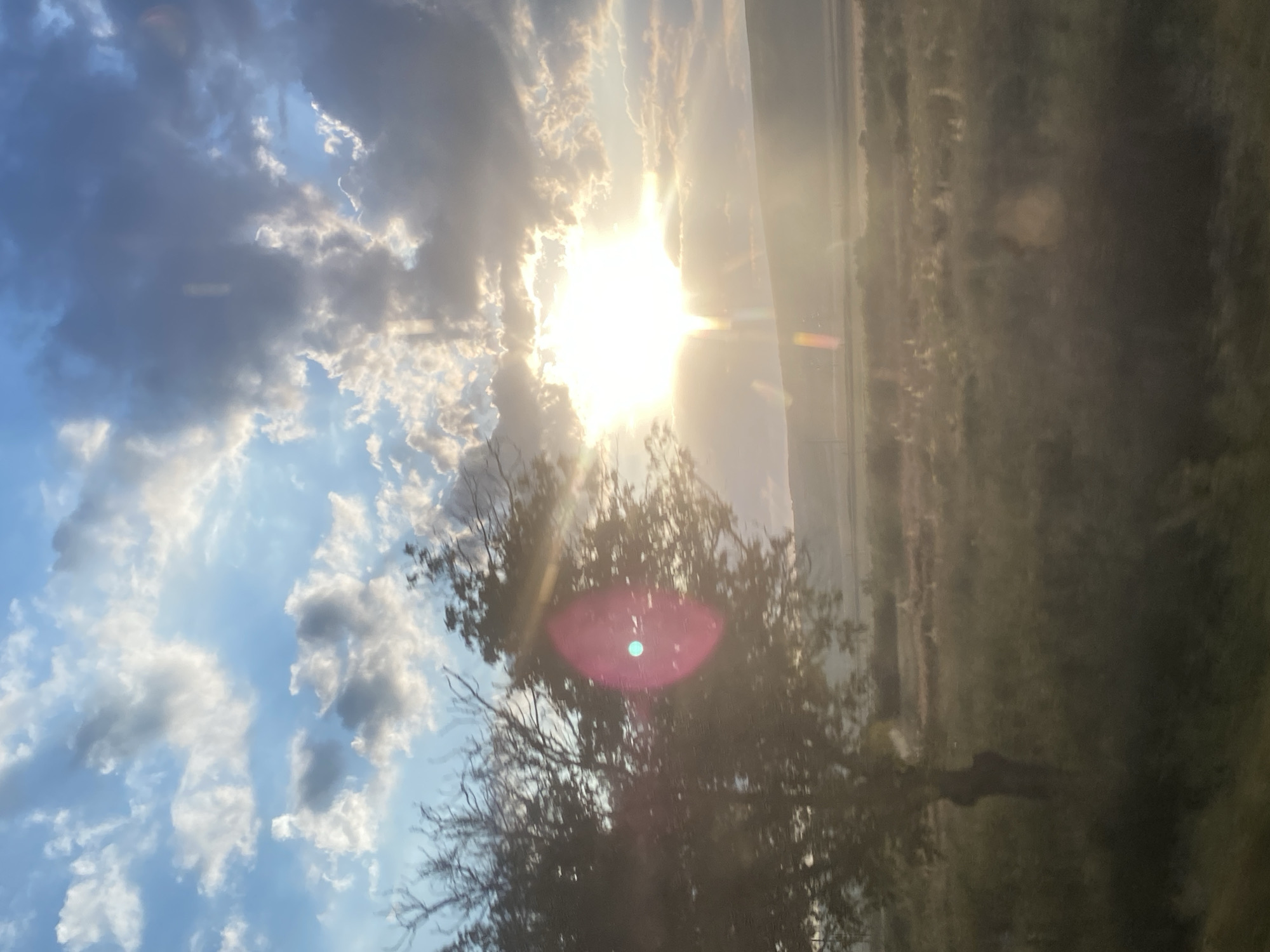 The image depicts a natural landscape under a dramatic sky. The sun is prominently featured, breaking through a combination of bright white and dark grey clouds, casting a warm, hazy light over the scene. Rays of sunlight emanate outwards, creating a lens flare effect, with distinct vertical streaks of light and a pinkish-blue circular anomaly visible.
In the foreground, to the left, a silhouette of a deciduous tree is visible, its branches bare against the sky. The tree appears to be situated in a grassy or shrub-filled area. Behind the tree, the terrain slopes gently upwards, forming a distant, muted hill. The middle ground is characterized by fields or meadows, with vegetation that appears dry and golden in the low sunlight. The overall impression is one of a rural or natural setting, possibly during sunset or sunrise, with atmospheric conditions contributing to the visual effect.
While the prompt asks for the location in Congaz, Moldova, there are no specific visual cues within the image that definitively identify it as such. The landscape is generic enough to be found in many rural areas. Therefore, without external information, attributing it to Congaz, Moldova, is not possible based solely on the visual content.