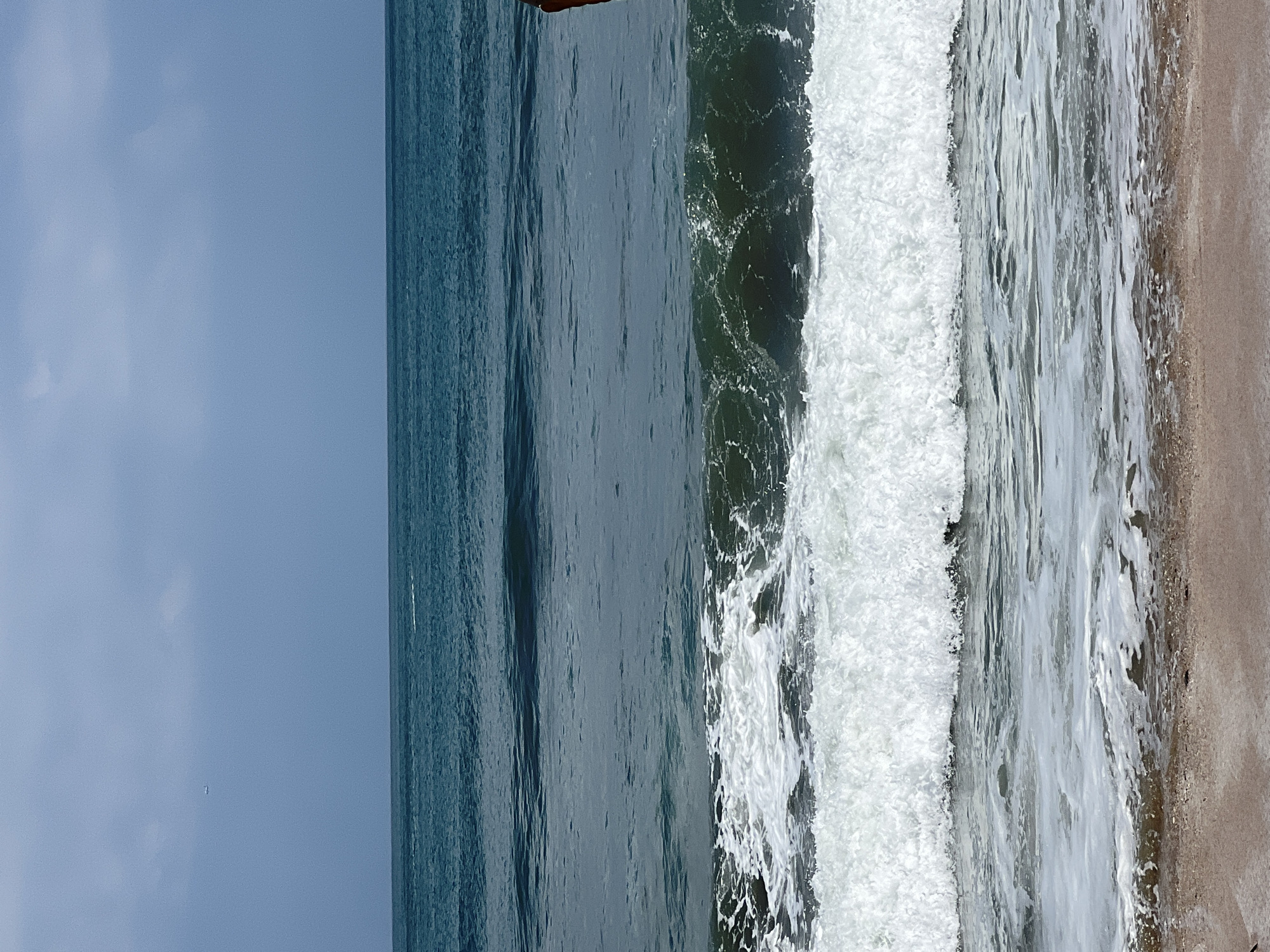 The image shows a beach scene in Kuta, Indonesia. The ocean is a deep blue, with waves crashing on the shore. The sky is a pale blue with some clouds. There are no people visible in the image. The scene is tranquil and serene.