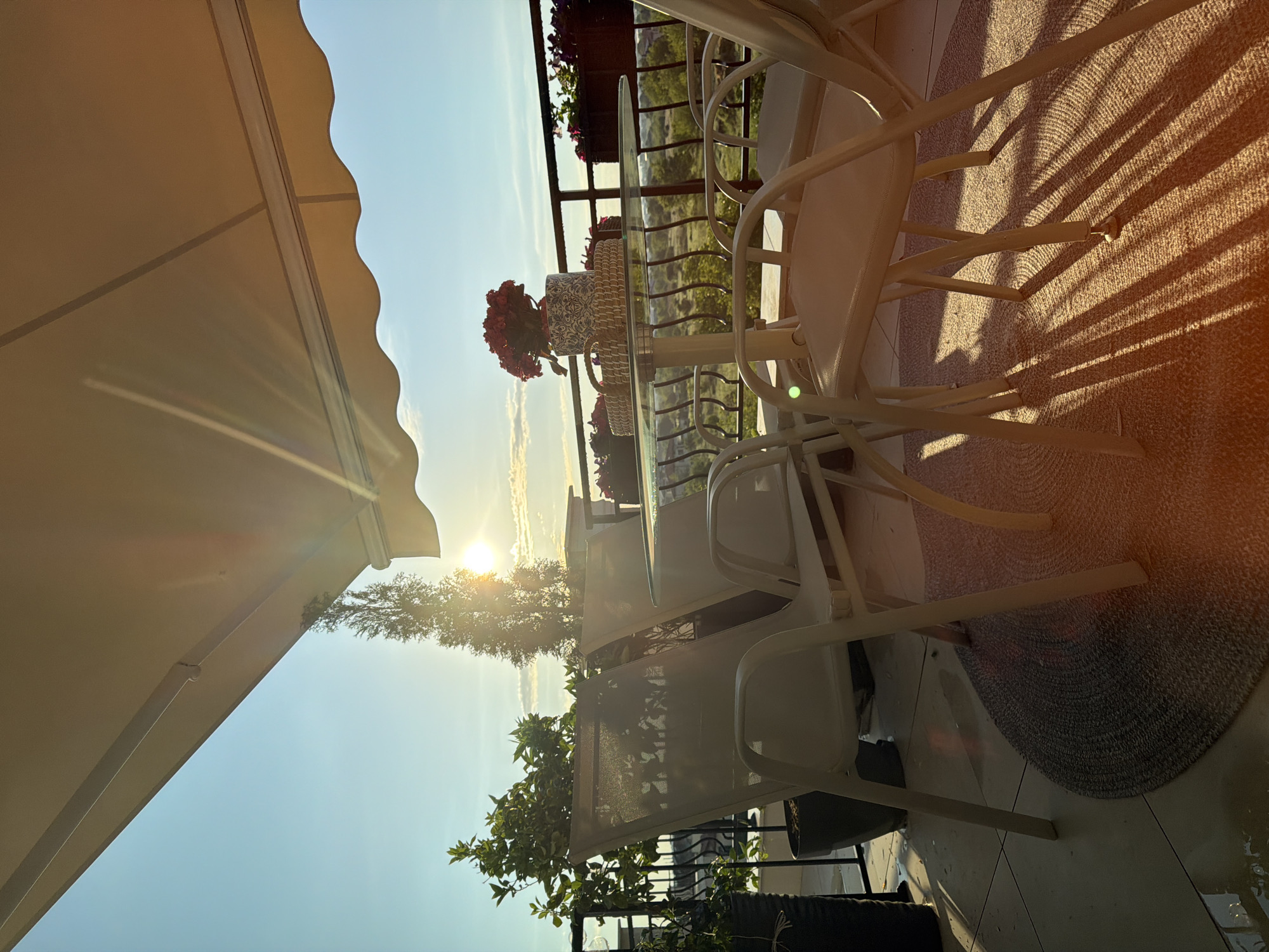The image shows a terrace in Plovdiv, Bulgaria, at sunset. There are white metal chairs around a glass table with a basket and a vase of red flowers. A beige awning covers part of the terrace. The terrace overlooks a landscape with trees and fields. There are flower boxes on the railing. The setting sun casts long shadows from the chairs onto the patterned rug. A small potted plant is in the foreground. The sky is blue with a bright sun.