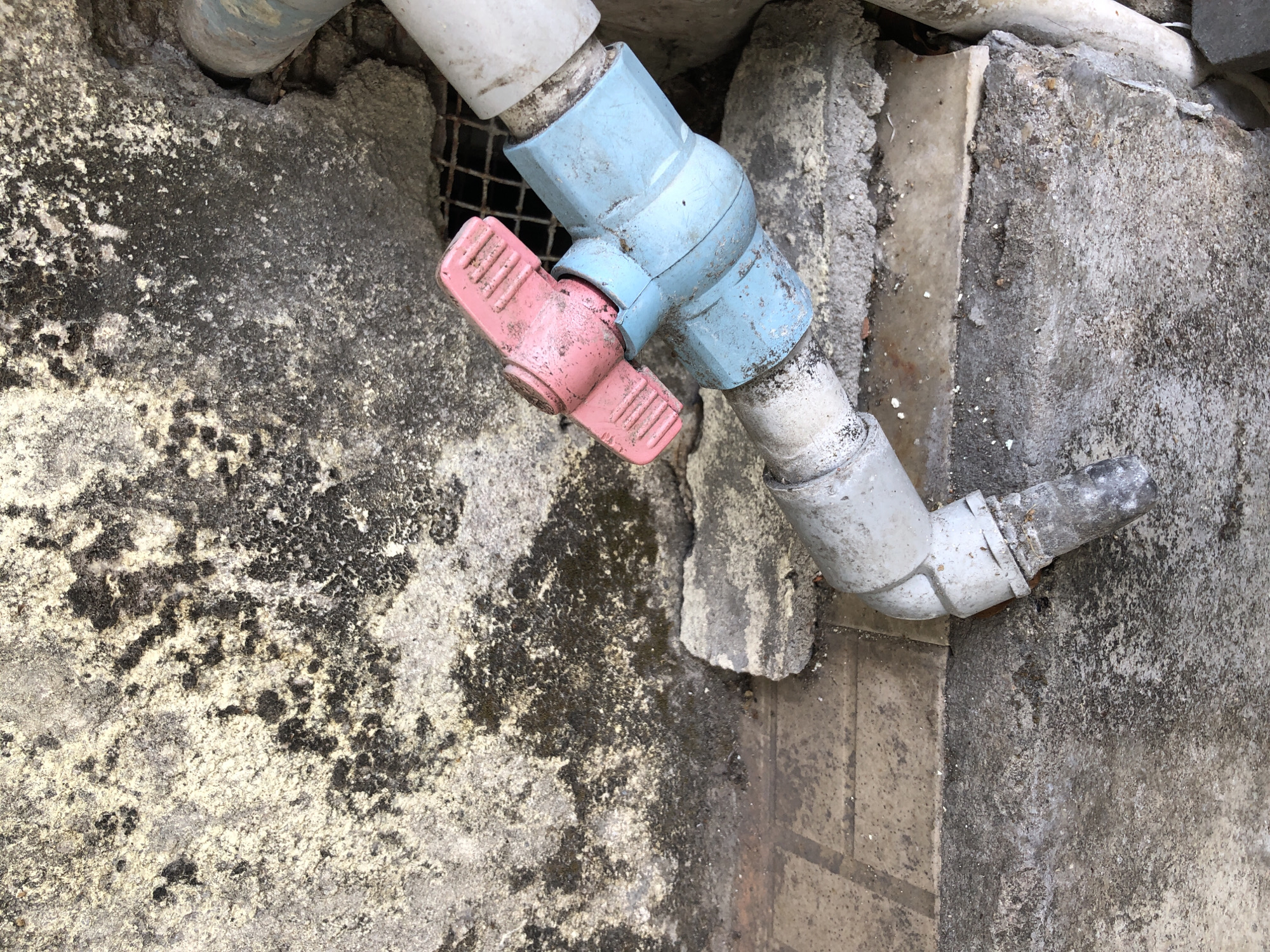 The image shows a close-up, high-angle view of plumbing fixtures and rough concrete surfaces. A pink handle controls a valve attached to a blue PVC pipe fitting, which connects to a white PVC pipe. A grey pipe with an elbow joint extends towards the bottom left of the frame. The background consists of textured concrete walls with areas of discoloration and some dark staining, possibly mold or grime. The ground appears to be made of cracked concrete slabs and dirt. The scene is outdoors and likely in a less maintained area, possibly an alley or utility space in Đà Nẵng, Vietnam. There are no people or animals visible. The lighting suggests it is daytime, with diffuse light casting soft shadows.