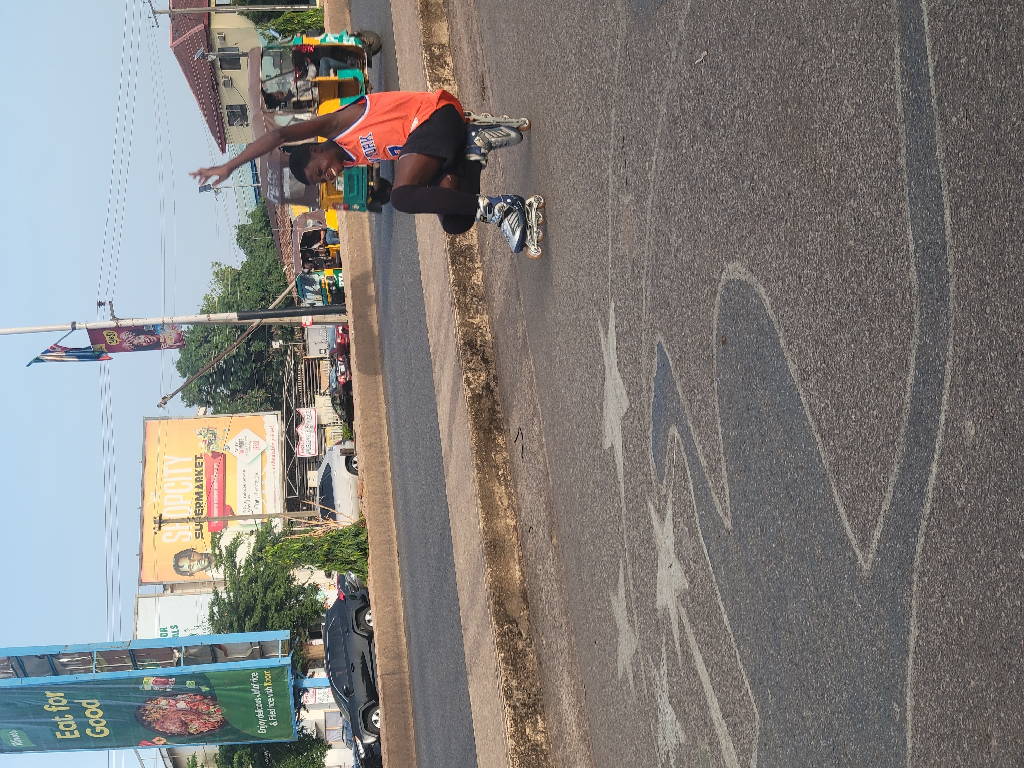 A young Black man is depicted inline skating on a paved street under a bright, clear sky. He is wearing an orange sleeveless jersey, likely a basketball uniform with "YORK" visible, black shorts, and black knee-high socks. He is in a dynamic pose, mid-stride, with his arms outstretched and a wide, joyful smile on his face, indicating enthusiasm and enjoyment in his activity.
The setting is an urban environment, identified by prominent billboards as Jos, Nigeria, specifically mentioning "NO. 35 Yakubu Gowon Way, Jos." The street surface is dark asphalt with some faded white and blue markings or graffiti. A concrete curb separates the main road from a rougher, unpaved strip. In the background, various vehicles, including colorful auto-rickshaws (tuk-tuks) and cars, are visible along with buildings and sparse greenery.
Notable details include the time of day, which appears to be sunny daylight. Emotions expressed by the subject are clearly joy and excitement. Visible text includes "YORK" on the skater's jersey, and on billboards: "SHOPCITY SUPERMARKET" with slogans like "WHY PAY MORE? when you can LESS! unbeatable prices!", and "Knorr Eat for Good" promoting "delicious Jollof rice & Fried rice."