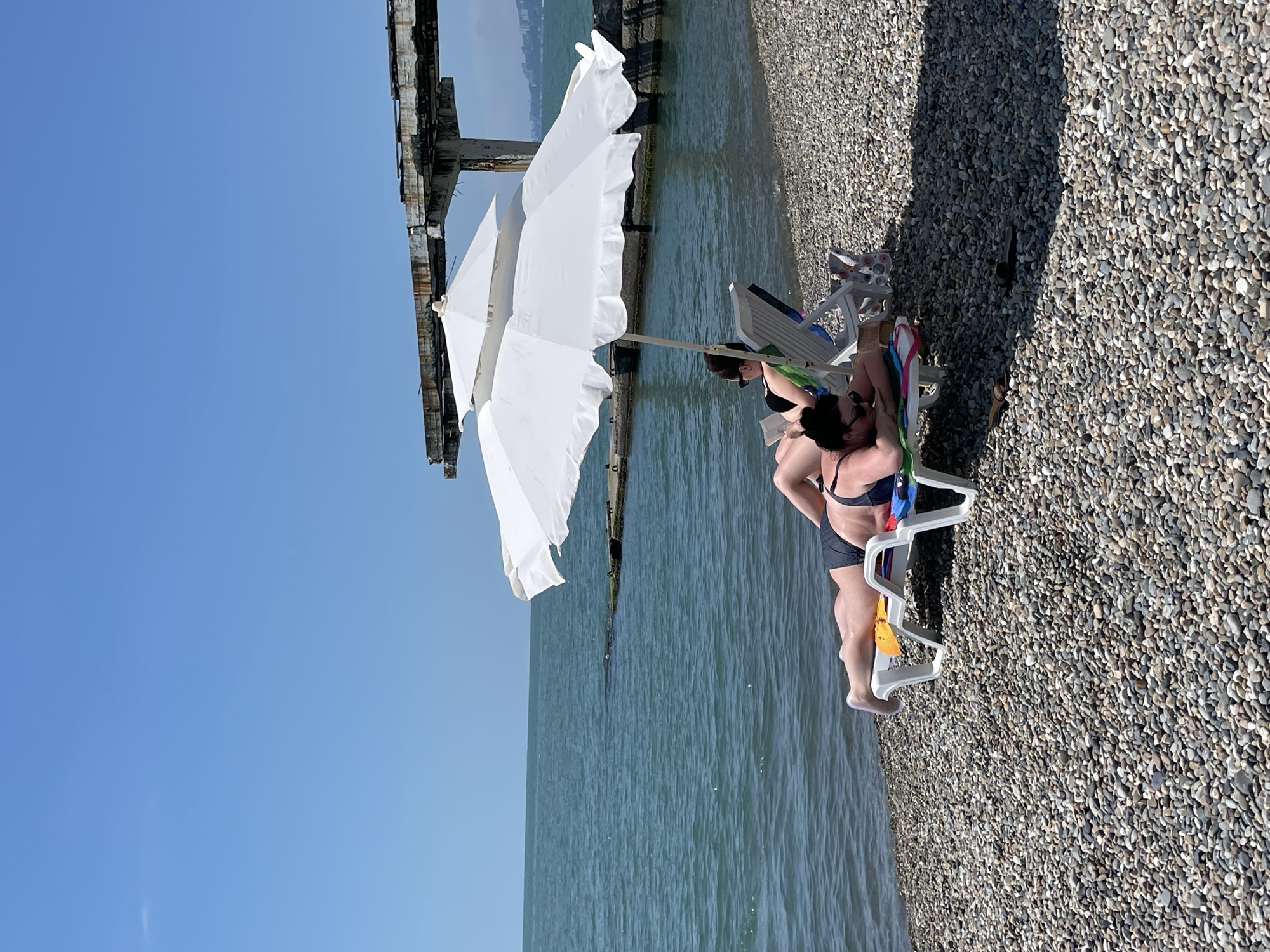 A bright, sunny day at a pebble beach in Sochi, Russia, showcases two individuals relaxing on lounge chairs under a white umbrella. The person in the foreground, lying on their stomach on a white lounge chair with a colorful striped towel, is wearing a dark bikini. Their legs are extended, and their body is oriented towards the camera. Behind them, another individual is reclined on a blue and white lounge chair, also wearing a dark bikini, with their head propped up, seemingly reading a book. A second, smaller white umbrella is positioned above and slightly behind the first one. The sea, with its blue-green water, is visible behind the beach, with a partially visible ruined pier or structure in the background extending into the water. The beach itself is covered in smooth, grey and tan pebbles. The sky is a clear, vibrant blue, with no clouds visible. The overall scene conveys a sense of leisure and relaxation on a warm summer day.