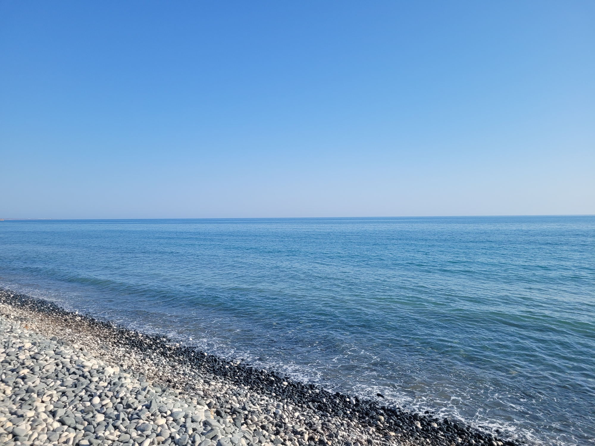 The image depicts a clear blue sky meeting a tranquil blue sea at a pebble beach. The foreground is dominated by a densely packed arrangement of smooth, grey and white pebbles, with a scattering of darker, almost black pebbles forming a distinct line where they meet the water's edge. The sea exhibits gentle ripples and the faint white foam of small waves lapping against the shore. In the distance, a faint line suggests the horizon where the sea meets the sky. The scene is devoid of any human presence, man-made structures, or vegetation. The overall impression is one of natural, undisturbed coastal serenity in Episkopi, Cyprus.