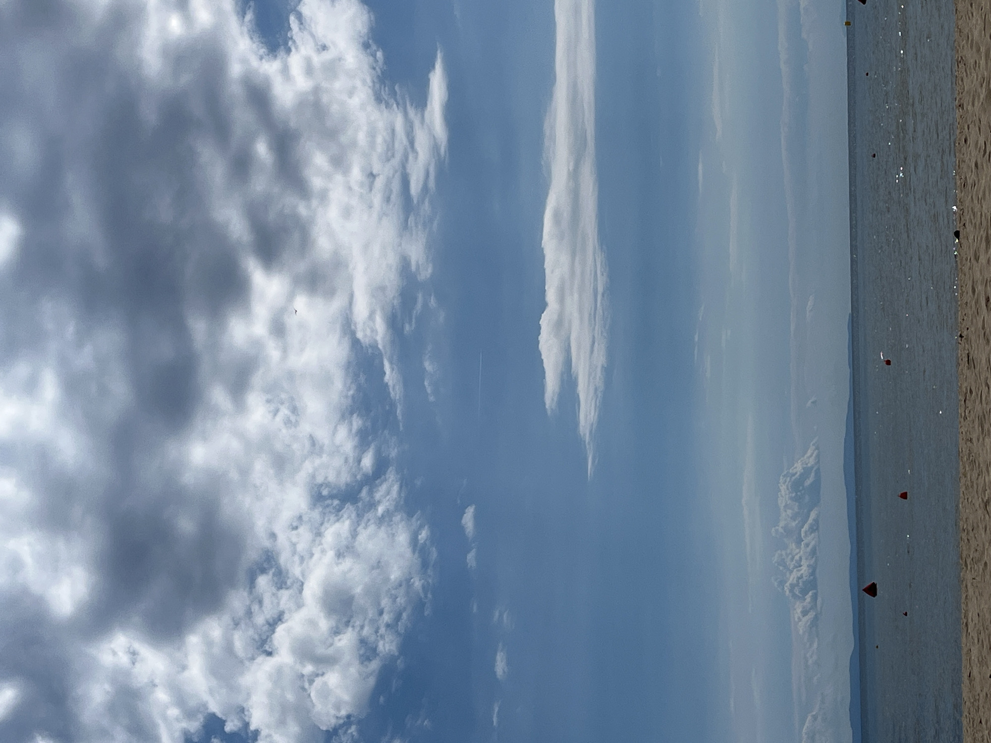 An overcast sky with scattered clouds looms above a calm sea. In the distance, a faint landmass with mountains is visible. The water is dotted with several orange buoys, and a sandy beach occupies the foreground. The scene is devoid of people.