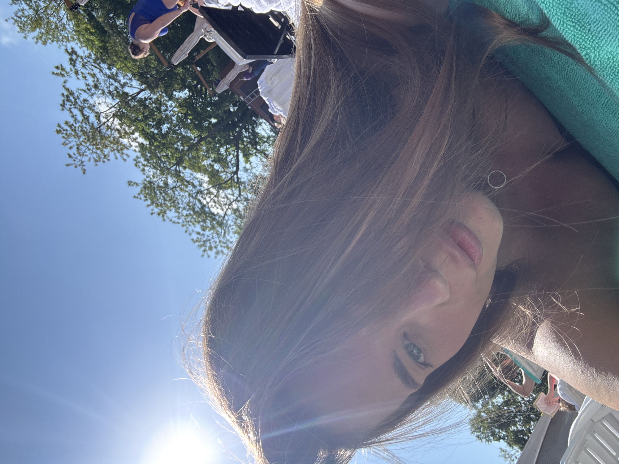 The image shows a young woman with light brown hair and blue eyes, likely taking a selfie outdoors. She is wearing a green top and a delicate necklace with a small circular pendant. The background includes a bright blue sky with the sun low in the frame, causing lens flare. In the distance, there are trees and glimpses of people gathered around what appears to be a table or structure. The location is suggested to be Sosnovy, Russia.
