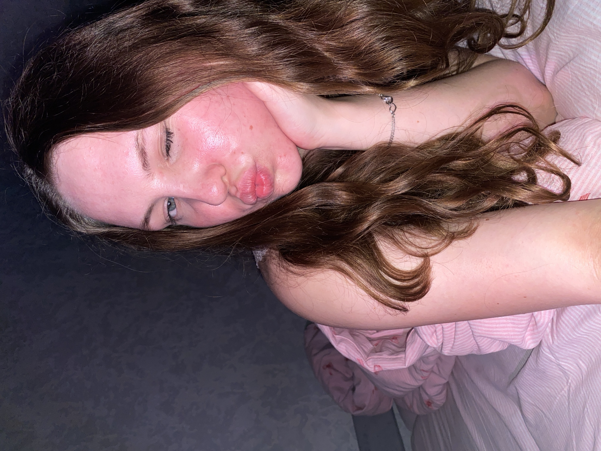 A young woman with long, wavy brown hair is lying on a bed in a dimly lit room. She is wearing a pink and white striped blanket. Her head is propped up on her hand, and she is winking and puckering her lips. Her skin appears red and flushed. The background is a dark, textured wall. There is no identifiable information to suggest this image is located in Izhevsk, Russia.