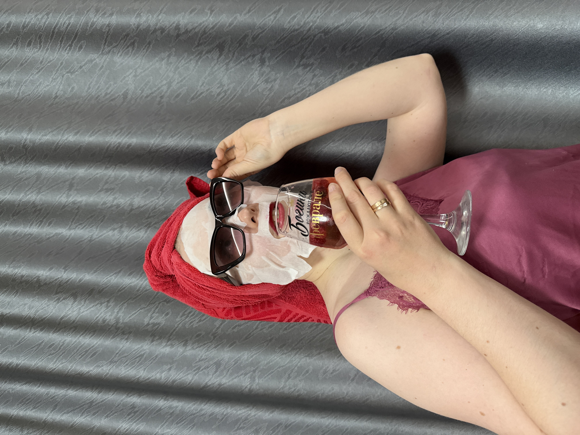A person is depicted from an overhead angle, reclining on a textured grey surface, likely a bed or sofa. They are engaged in a self-care routine, wearing a white sheet mask that covers most of their face, accented by oversized black sunglasses. A red towel is wrapped around their head, covering their hair.
The individual is dressed in a burgundy or maroon silk-like camisole or robe, with visible lace detailing from a matching bralette or underwear underneath. Their left arm is bent, resting near their head, while their right hand holds a large, clear stemmed glass filled with a reddish beverage that appears to contain small fruit pieces or ice. Two gold-toned rings are visible on the ring finger of their right hand.
Visible text on the glass is in Cyrillic, with "БОРЩИ" (Borshch) clearly readable at the top of the label, and below it, partially obscured, "рождаются в КРАЛЕ" (rozhdayutsya v KRALE), translating to "Borscht is born in KRALE." The overall scene conveys a sense of relaxation and indulgence in an indoor setting. The lighting is even, suggesting an indoor environment without strong external light sources.