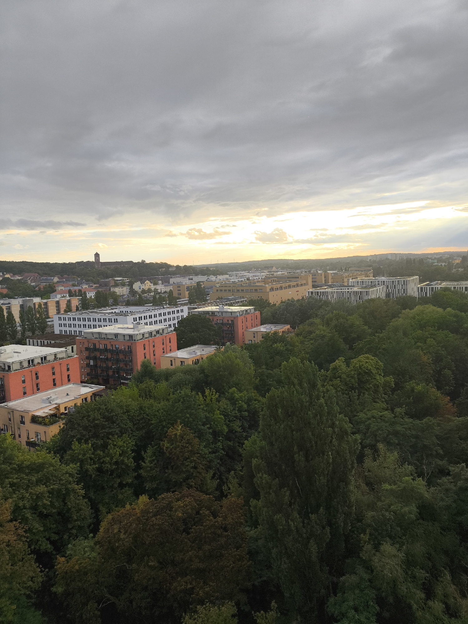 This is an aerial, high-angle, vertical shot taken from an elevated perspective, looking out over a cityscape characterized by a blend of modern apartment buildings, dense greenery, and a hazy sky. The image captures a portion of Potsdam, Germany, specifically showing residential buildings interspersed with mature trees and parkland.
In the foreground, a lush canopy of trees dominates the lower half of the frame, with various shades of green indicating different species and densities of foliage. Some of the trees are tall and slender, while others are broader and more spread out.
Rising above the trees, to the left and center, are several multi-story apartment buildings. Some are painted in vibrant hues of orange and red, featuring visible balconies and windows. Other buildings are more contemporary in design, with light-colored facades and linear architectural details. One particularly striking building in the mid-ground has a white exterior with rows of dark windows and green horizontal accents on its roof.
In the distance, the cityscape extends, with more buildings visible, some appearing lighter in color and more uniform in design, suggesting office or commercial structures. A tall, dark tower-like structure is visible on a hill in the far distance, possibly a historical landmark or a communication tower. The horizon is softened by a hazy, overcast sky, with patches of pale sunlight breaking through the clouds, creating a diffused, soft lighting effect. The overall impression is one of urban development harmoniously integrated with natural landscapes. There are no visible people in the image.