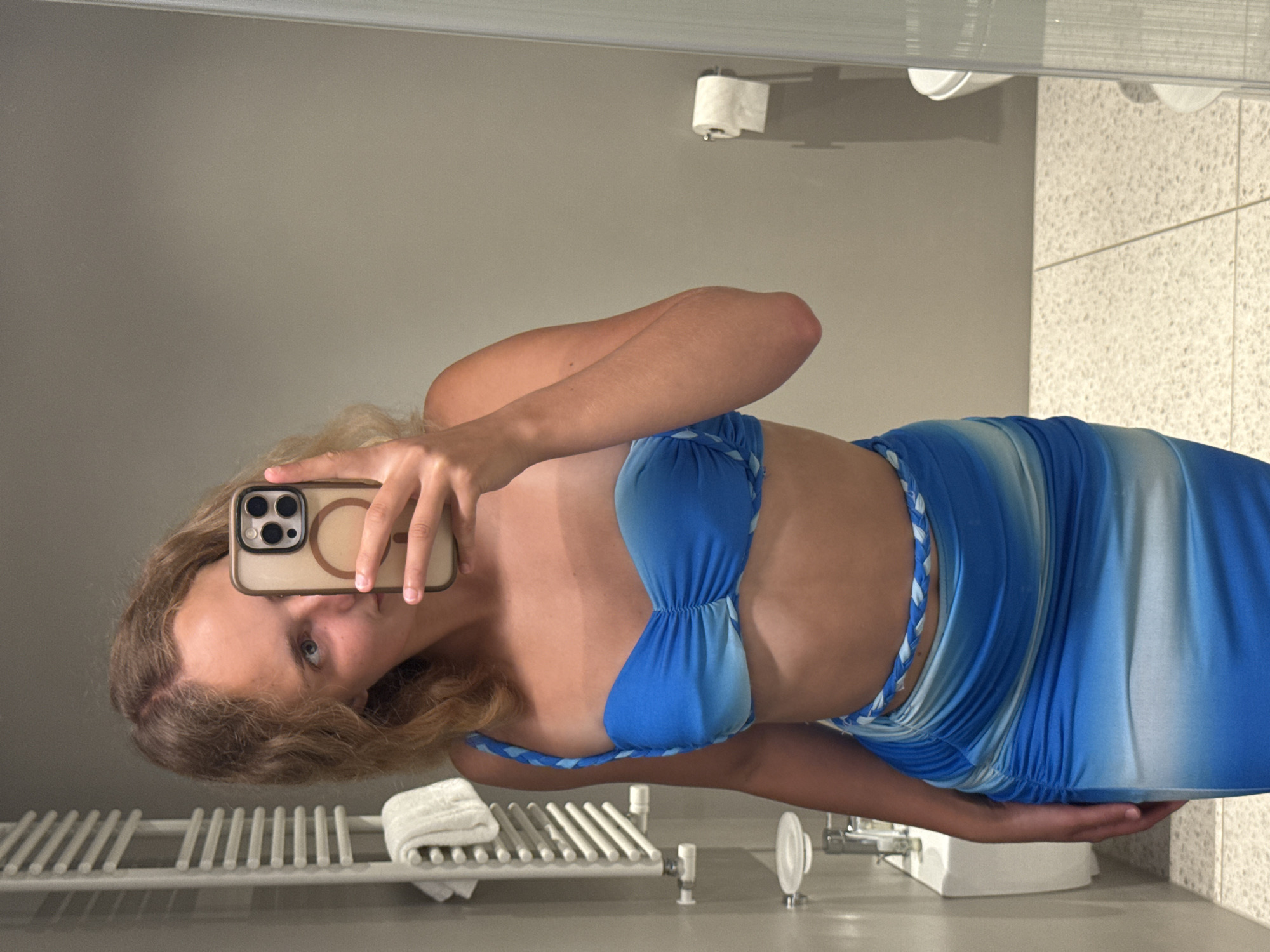 A young woman with blonde hair is taking a mirror selfie in a bathroom. She is wearing a blue and white tie-dye bikini top and a matching ruched maxi skirt with a braided rope belt. The bathroom has a gray tiled wall, a white radiator with a towel on it, a soap dispenser, and a toilet paper holder. The floor is a light-colored terrazzo. The image is captured in a hotel bathroom, likely in Trieste, Italy, given the context. The overall impression is of a vacation setting, possibly a hotel room.
