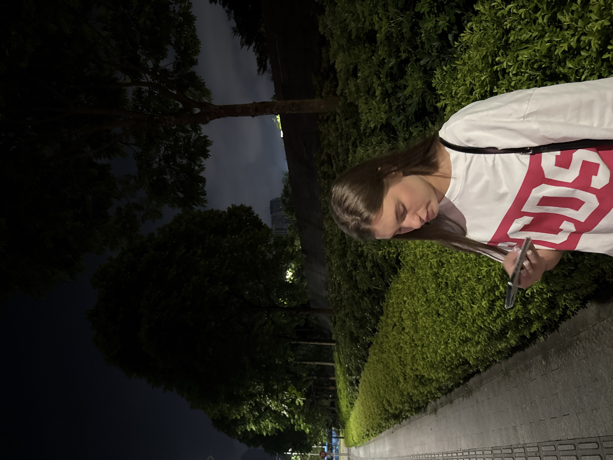A young woman is viewed from the side as she looks down at a smartphone she is holding. She has long, brown hair and is wearing a white t-shirt with a large, red graphic design. The background consists of lush green bushes and trees, with a paved walkway in the foreground. The scene is dimly lit, suggesting it is nighttime, and the sky is dark and cloudy. A streetlamp can be seen in the distance, illuminating part of the walkway and the surrounding foliage. The overall impression is one of a quiet, urban setting at night, possibly a park or a residential area.