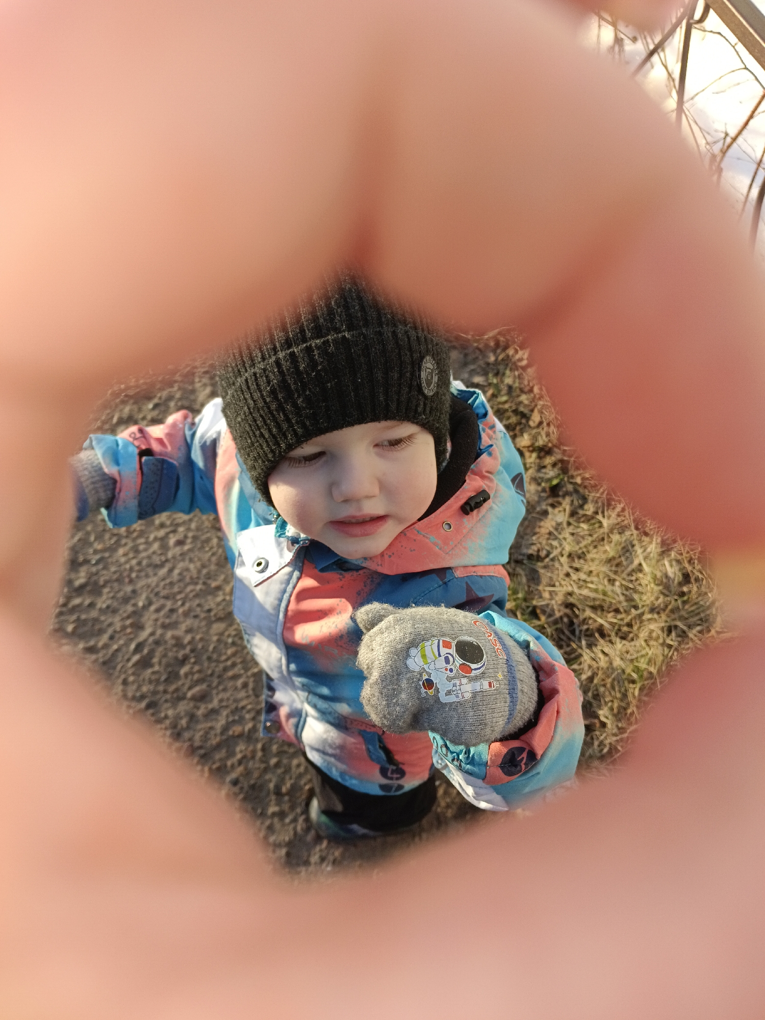 A young child stands outdoors in Chaikovsky, Russia. The child wears a black knit cap and a blue, orange, and white winter coat. The child's left hand is visible, covered by a gray mitten with a cartoon astronaut design. The child is framed by blurred human fingers. The ground appears to be a combination of asphalt and dormant grass. Bare branches and a fence are visible in the upper right corner of the frame.