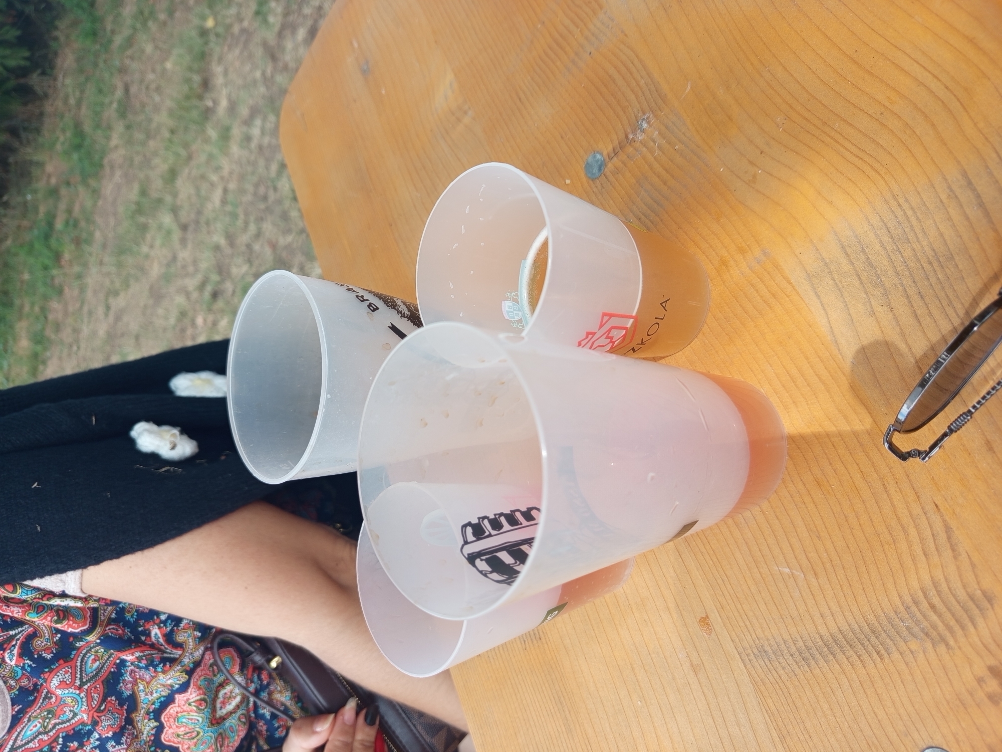 Four translucent plastic cups containing beverage are arranged on a light brown wooden table outdoors. One cup, closer to the foreground on the right, contains a golden-colored liquid with a frothy head, and has "ZKOŁA" printed on it in red. Another cup, to its left, is partially filled with a pinkish liquid and has a black and white illustration of a microphone inside. A third cup, further to the left, also contains a pinkish liquid. The fourth cup, on the far right and behind the others, is empty and has the word "BRA" visible on its side. The background shows green grass and foliage, indicating an outdoor setting. In the upper left, a person's arm wearing a colorful patterned shirt and a black cardigan is visible, along with a dark handbag. The lighting suggests it is daytime.