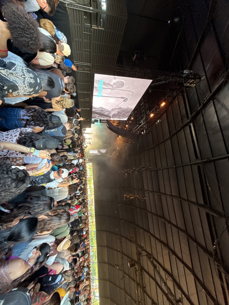 The image captures a densely packed crowd at a concert in what appears to be a large, covered venue. The image is taken from an elevated perspective looking down and forward towards the stage, likely near the front of the crowd. The venue has a high, arched ceiling with visible support structures and rigging for lights and stage equipment. A large screen displays a black and white image. There is smoke or haze rising towards the stage area. The location is Indio, United States.