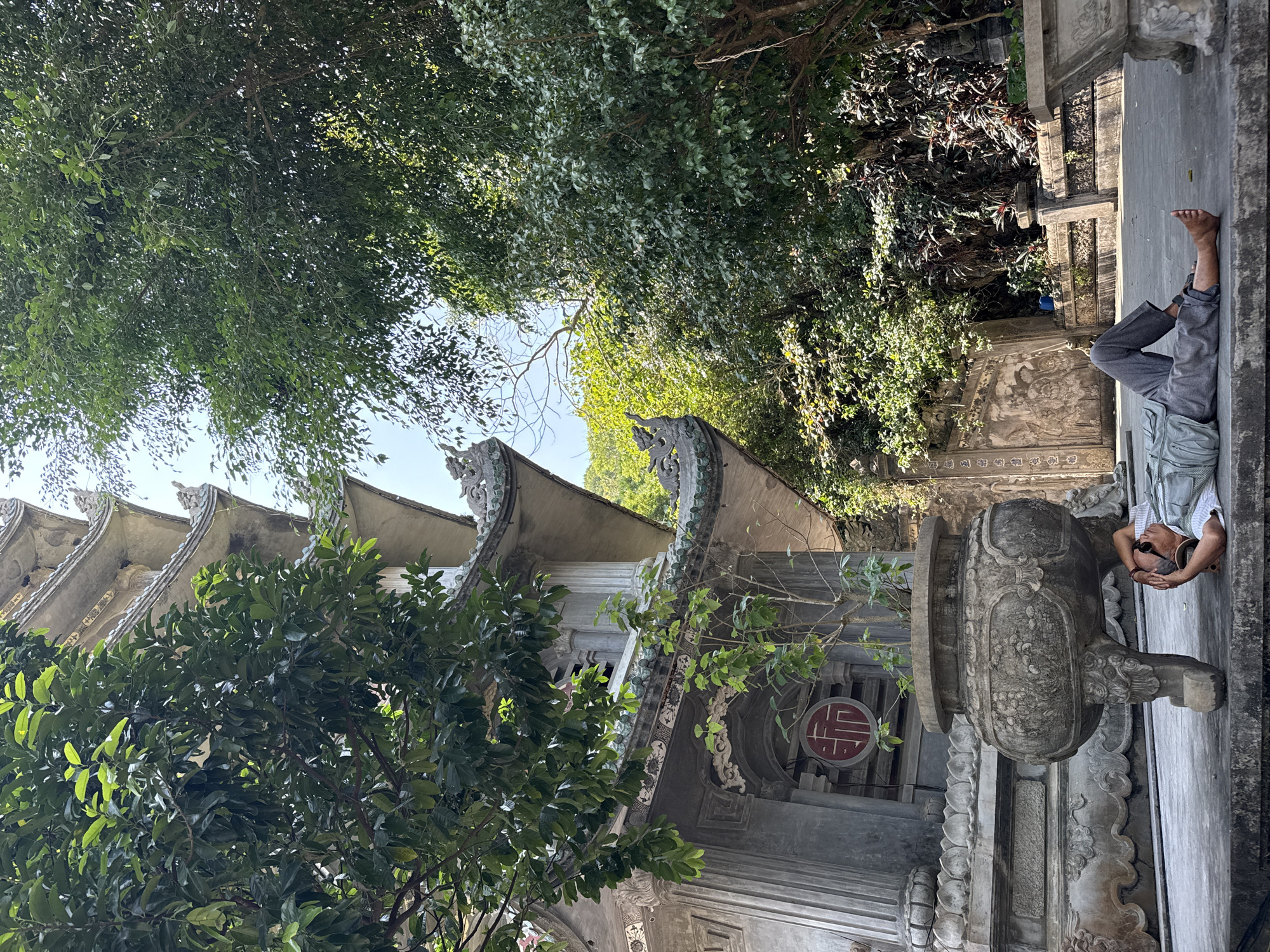 The image depicts an outdoor scene at a pagoda complex, identified as being within the Marble Mountains of Đà Nẵng, Vietnam. In the foreground, a single adult male lies supine on a light grey concrete platform. He is barefoot, dressed in grey trousers and a light-colored, possibly striped, short-sleeved shirt, with dark sunglasses resting on his forehead. His hands are clasped behind his head, and his eyes appear to be closed, indicating a resting posture.
To the left of the man, a large, ornate grey stone urn or incense burner stands on four stylized, carved legs. The urn features intricate bas-relief carvings depicting floral and faunal motifs, and a small green plant grows within its opening. Behind the urn and the resting man, the lower tiers of a multi-level concrete pagoda structure are visible on the left side of the frame. This section features decorative columns, grey relief carvings, and a prominent red circular emblem containing a white character, likely "Phúc" (福), representing "good fortune." The pagoda's rooflines exhibit curved, tiled sections with additional decorative elements, some resembling mythical creatures.
Dense green foliage, comprising numerous trees and undergrowth, occupies the right side and background of the image, partially obscuring the pagoda and revealing sections of a bright, clear sky above. A concrete wall with further bas-relief carvings is discernible behind the man on the right. The scene is illuminated by natural daylight, casting some shadows.
