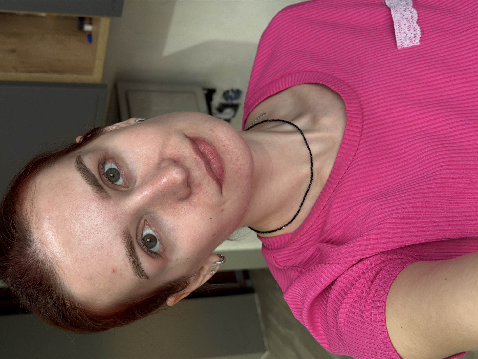 A close-up selfie shows a woman with light skin, auburn hair pulled back, wearing a bright pink ribbed shirt with a small white lace accent near the shoulder. She has green eyes, noticeable freckles, and is wearing small earrings. A thin black beaded necklace adorns her neck. The background shows a partially visible grey and light brown cabinet. The specific location within Chaikovsky, Russia, is not identifiable from the provided image.