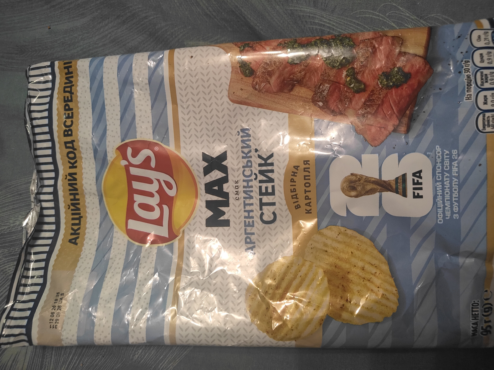 This is a close-up, top-down shot of a bag of Lay's potato chips. The bag features a patriotic Ukrainian design with blue and yellow stripes, and images of steak and potato chips. Text on the bag indicates it is a promotional item, possibly related to the FIFA World Cup, and lists nutritional information. The bag is placed on a blue, textured surface, likely a piece of fabric. The lighting suggests an indoor setting, and the overall mood is casual and consumer-oriented. The presence of Ukrainian text on the packaging confirms the location context.