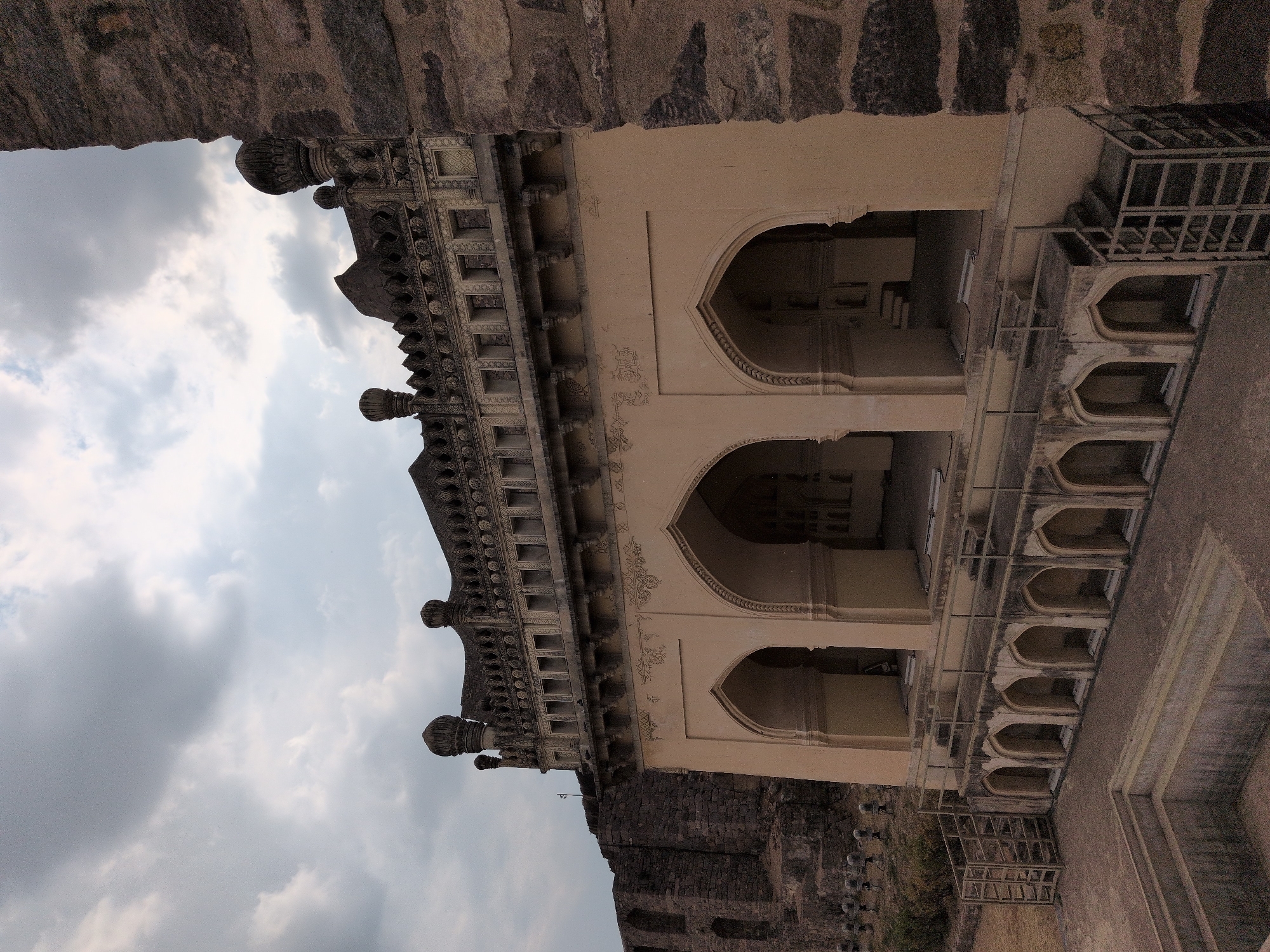 This image depicts a historic architectural structure, likely a part of a fort or palace complex, situated under an overcast sky in Hyderabad, India. The primary subject is a multi-tiered building constructed of stone and stucco.
The building's lower level features three large, pointed arches forming open bays, set into walls of a light beige or ochre color, adorned with subtle decorative patterns above the arch spandrels. Below these arches, a series of smaller, recessed arched openings are visible, forming a base. A metal railing system with a small staircase is present along the perimeter of this lower level. The ground in the foreground is composed of dry, compacted earth or stone, and an empty, rectangular stone water tank or pool structure is prominently featured in the lower-left quadrant.
The upper level of the main building is characterized by a richly decorated parapet, featuring a repeating series of small, open-arched niches or windows, capped with ornate dome-shaped finials. This upper section is darker in tone, likely utilizing different stone types or finishes.
To the left of the main structure, extensive, rough-hewn dark stone walls, indicative of fortification, extend into the background, also featuring arched openings. A row of smaller, pillar-like stone objects is partially visible on the ground in this area. On the extreme right of the frame, a large, rugged stone wall, consistent with fort construction, forms a boundary. The sky above is entirely covered with dense, grey clouds, indicating an overcast condition. No individuals or animals are present in the scene. The overall impression is one of an ancient, weathered, and uninhabited historical site.